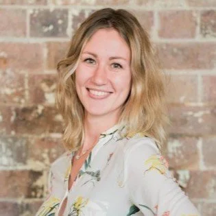 Portrait of a woman with blonde hair smiling, standing against a brick wall.