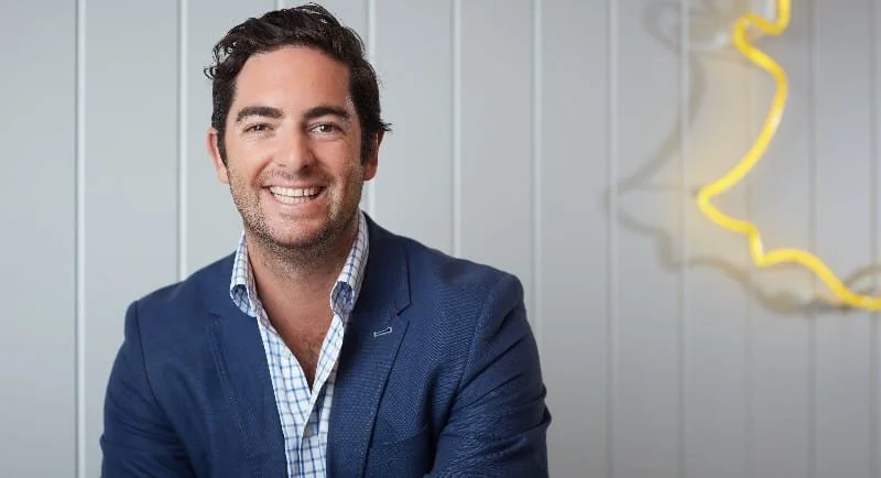Smiling man in a blue blazer and checkered shirt sitting in front of a light gray wall with a yellow neon light design.