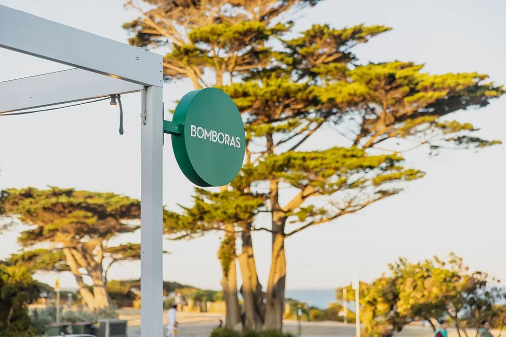 A green circular sign with the word 'BOMBORAS' mounted on a white metal structure, with tall trees in the background during daylight.