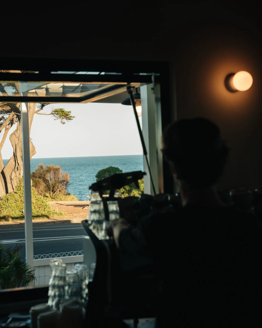 Person behind a coffee machine indoors, looking out a window at the ocean, with trees and a road visible outside.