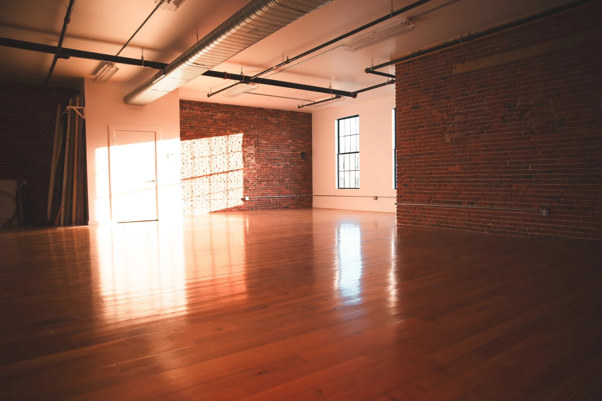 Baltimore photography studio with hardwood floors, exposed brick walls, and large windows. Sunlight casts shadows on the walls and floor, and the ceiling has exposed ductwork and lighting fixtures.