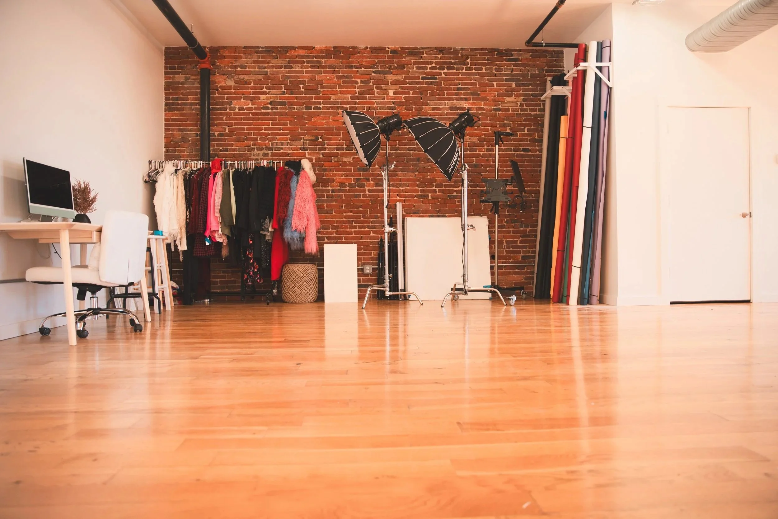 Empty photography studio with studio lighting, clothing rack with various costumes, and colorful backdrop rolls against an exposed brick wall.