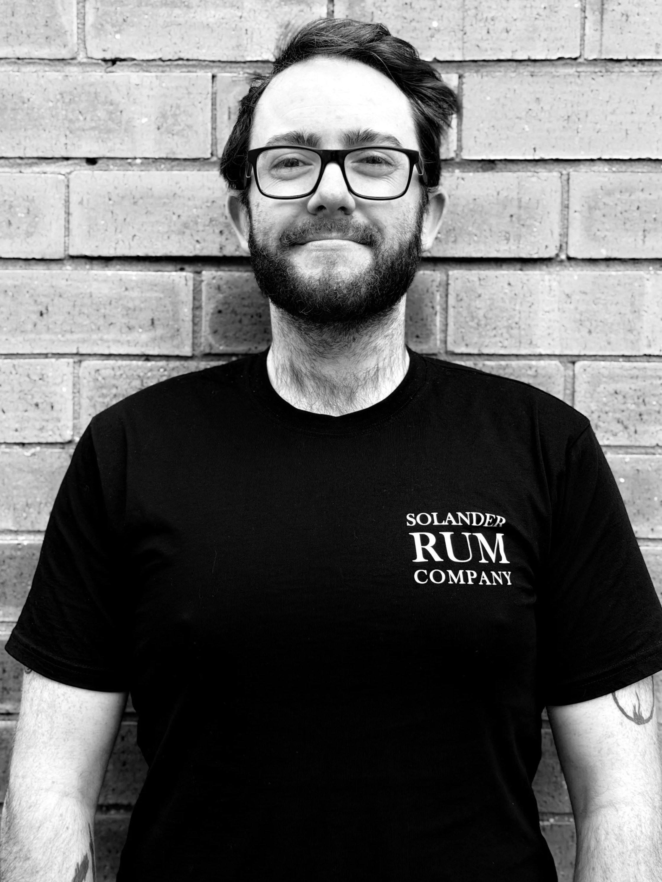 A smiling man with short hair, wearing a black shirt with a logo that reads 'Solander Rum Distillery,' standing outside against a brick.