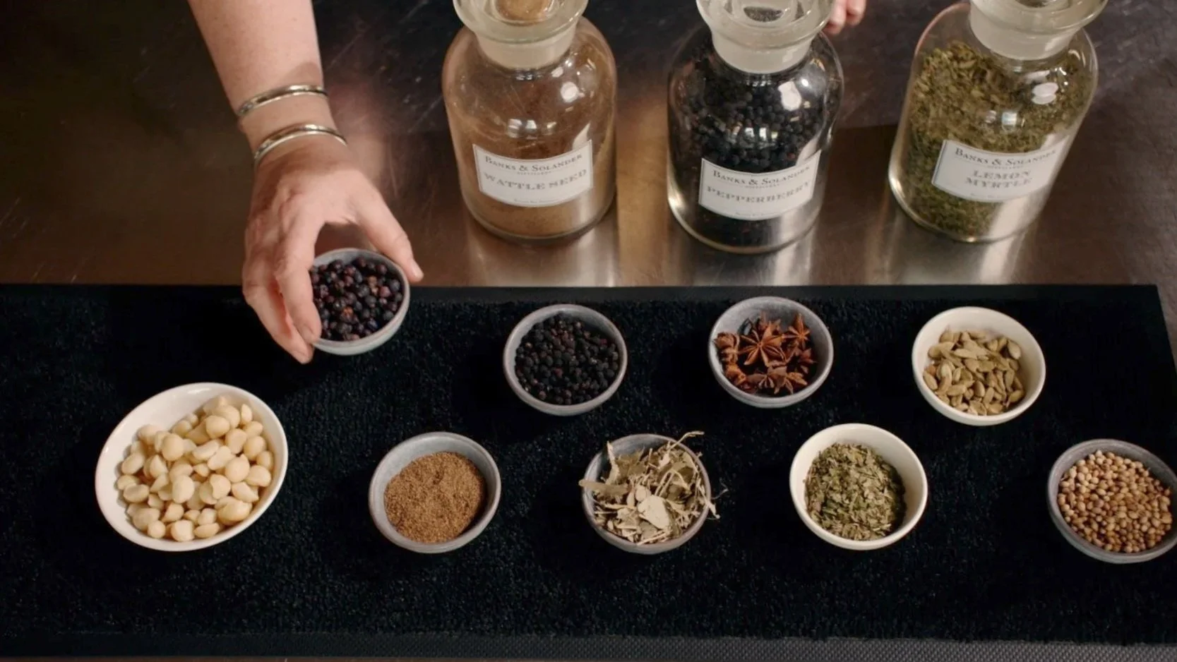 A person with a bracelet on their arm holding a small bowl of mixed dried berries, with various other dried herbs and spices in small bowls and jars arranged on a black surface.