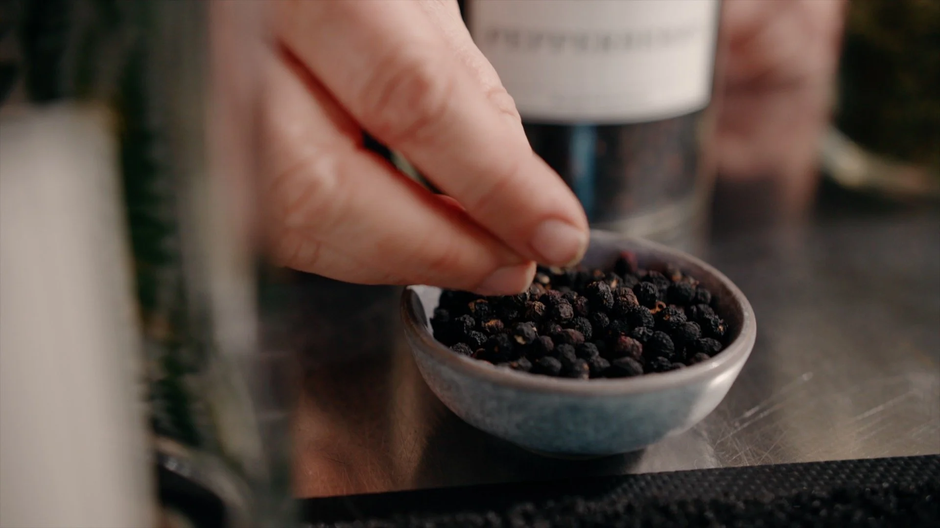Close-up of a hand holding black peppercorns above a small bowl of more peppercorns on a metal surface.