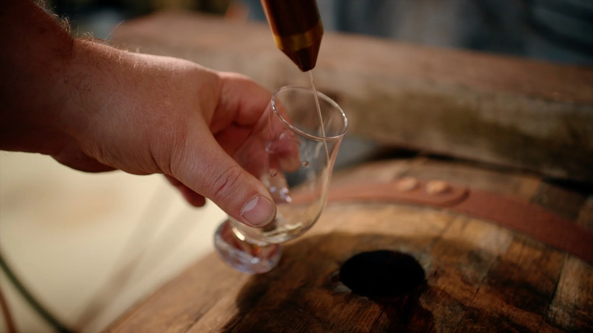 Person pouring whiskey into a small glass over a wooden surface, possibly a barrel.