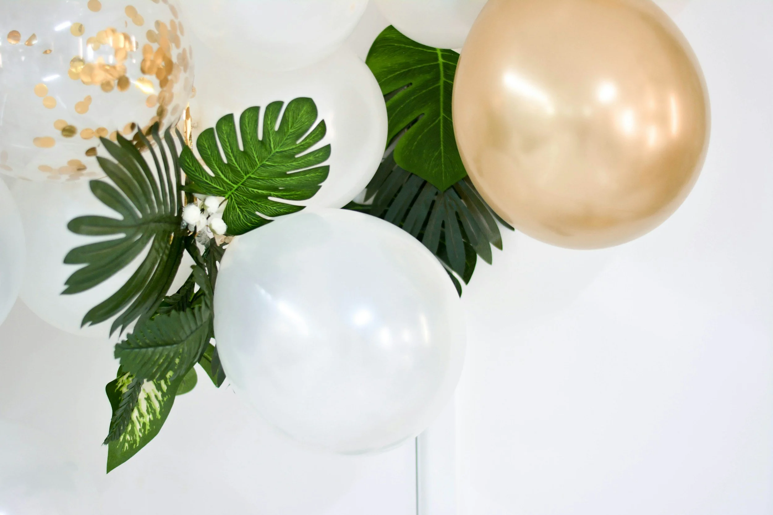 A decoration of white, gold, and transparent balloons intertwined with green tropical leaves.