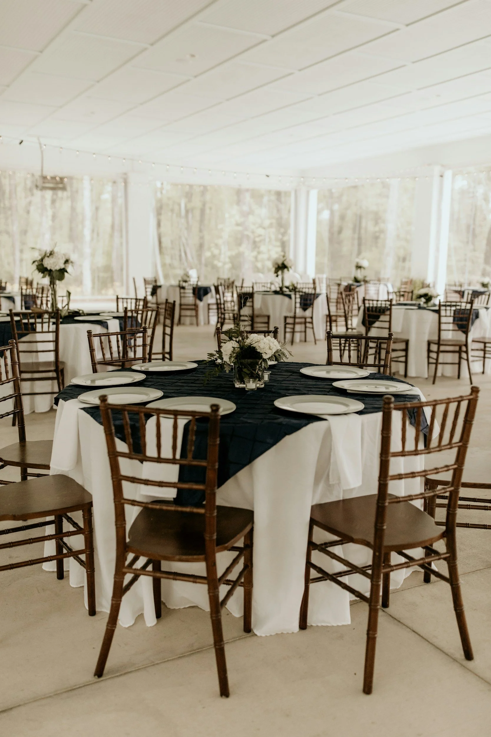Round banquet table with a white tablecloth and a black overlay, set with white plates, and a floral centerpiece, surrounded by wooden chairs, in an outdoor tent with string lights and trees visible in the background.