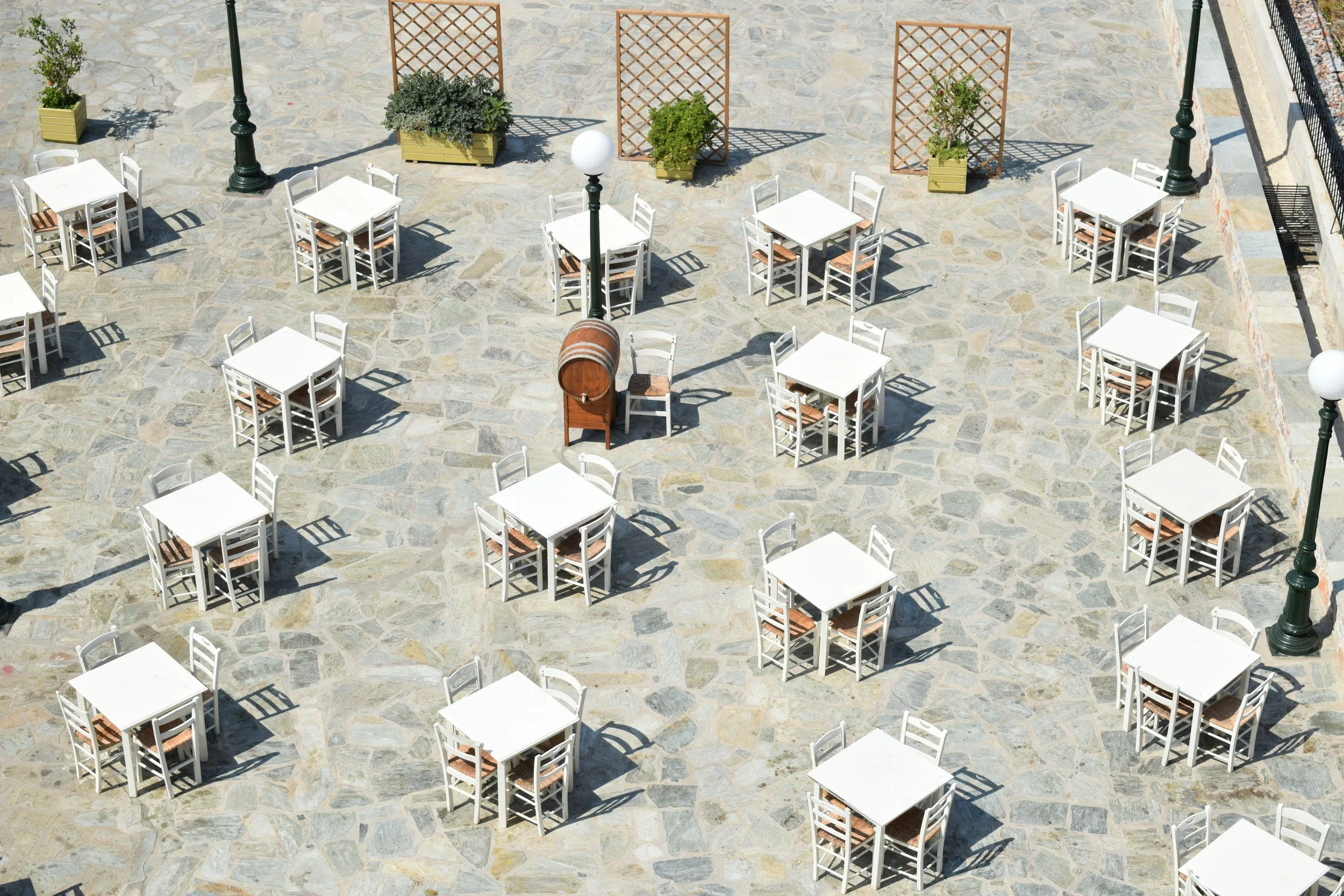 Empty outdoor patio with white tables and chairs, a wooden barrel, potted plants, large lamp posts, and decorative trellises on a stone ground.