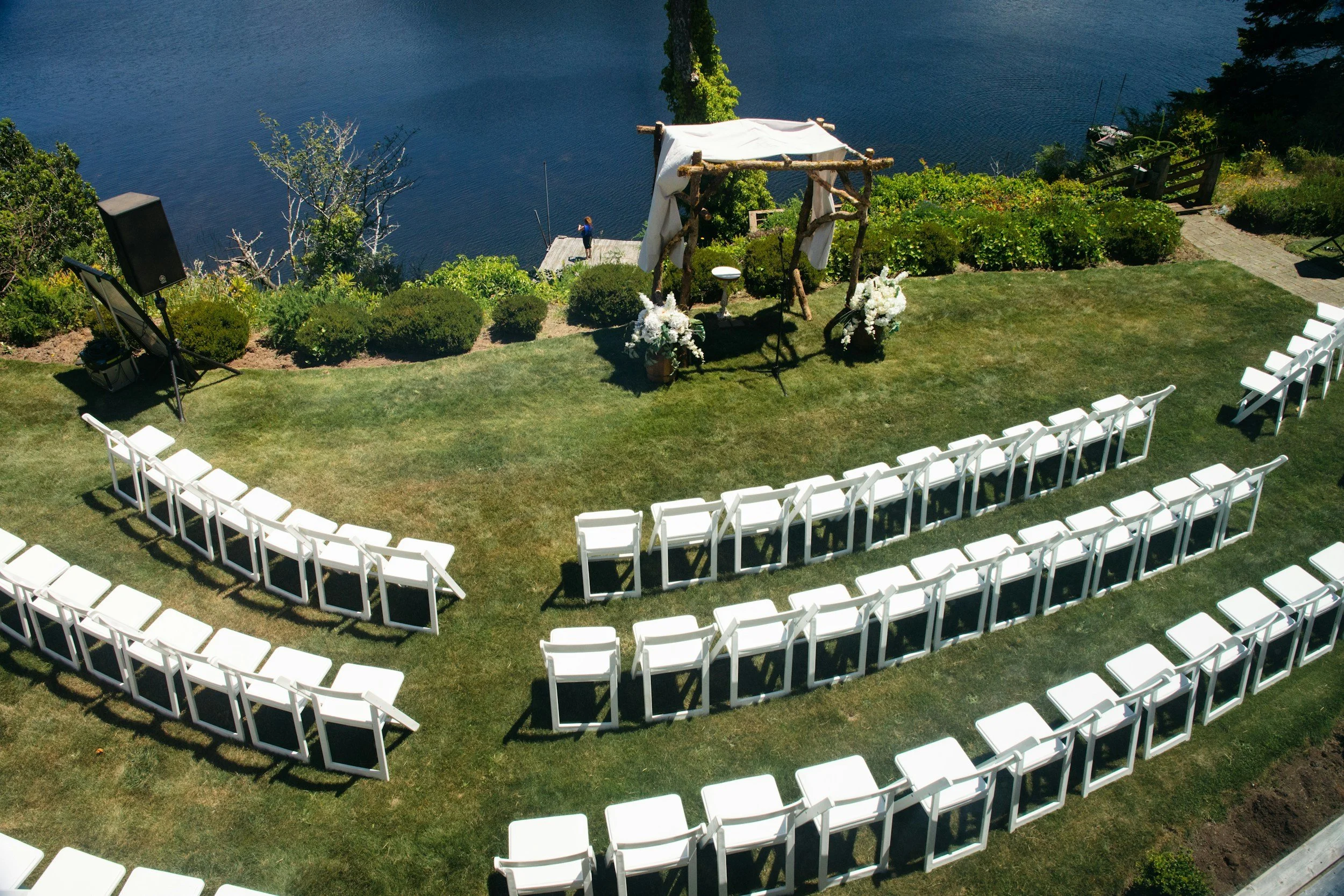 Outdoor wedding setup with white chairs arranged in a semi-circle, a small wooden arch decorated with white drapery and flowers, overlooking a lake, with greenery and a speaker on a stand nearby.