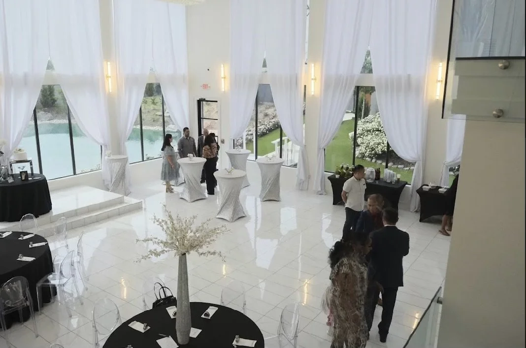 Guests mingling at a bright, elegant reception with white curtains, large windows, and decorated tables.