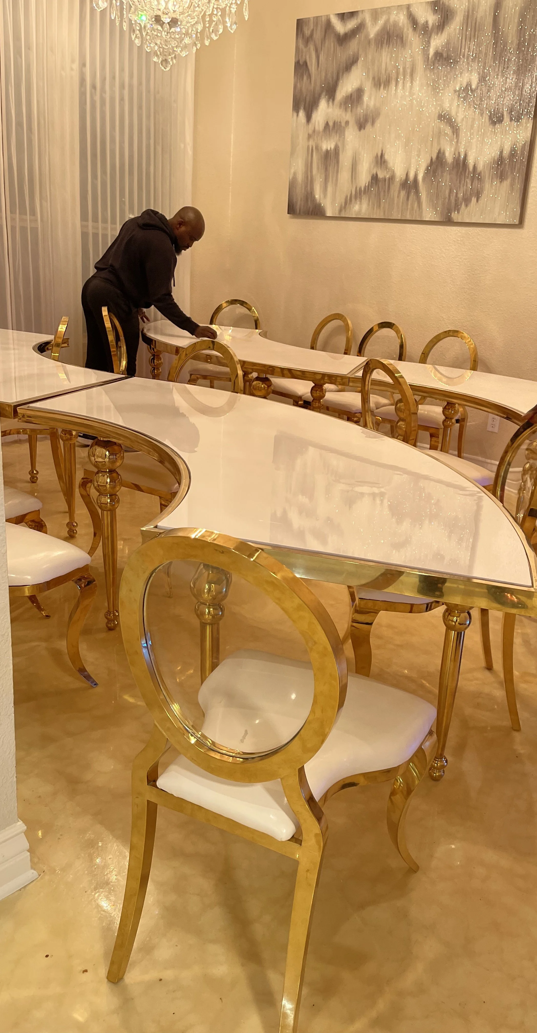 A man cleaning a luxurious gold and white dining table in a well-lit dining room with a chandelier and abstract wall art.