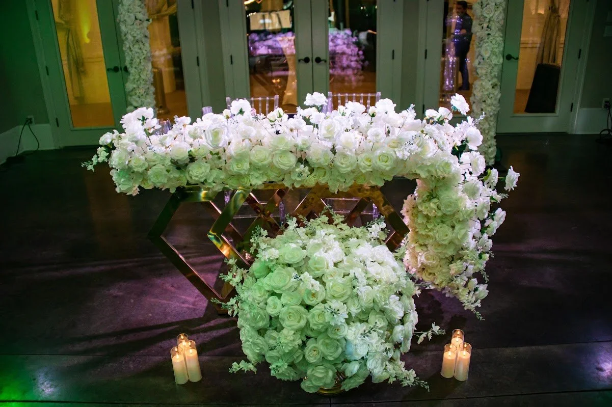 A floral wedding centerpiece with white roses and flowers on a metallic stand, illuminated by candles at the base, placed on dark wooden floor in front of glass double doors decorated with white flowers.
