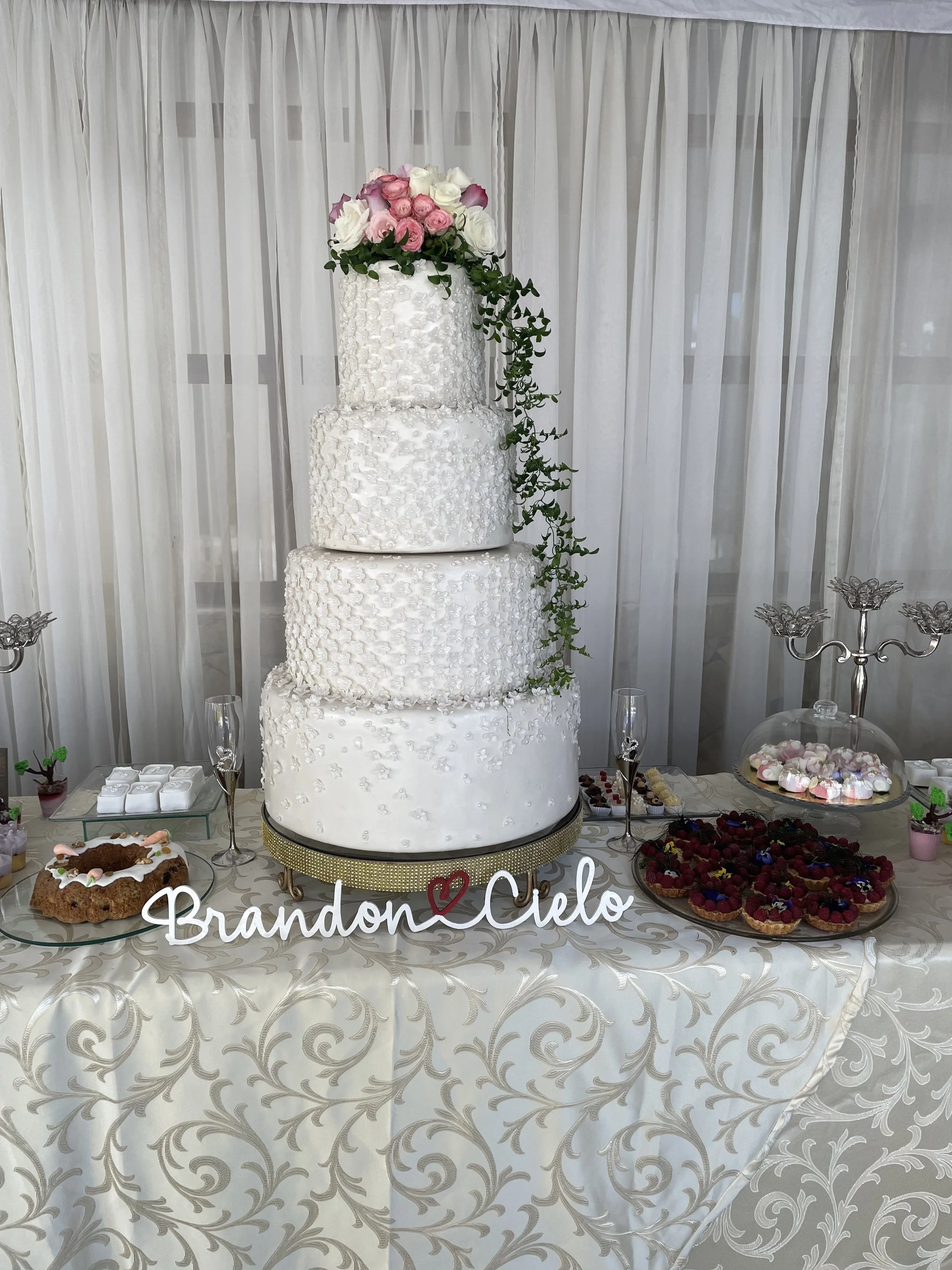 Four-tier white wedding cake with floral decorations on a table with desserts and a sign that reads "Brandon ♥️ Cielo".