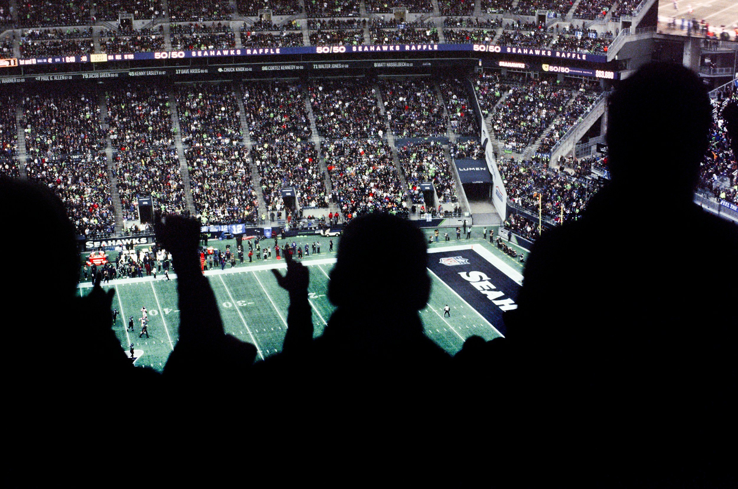 Silhouetted spectators watching a football game at the Seattle Seahawks stadium, with the field and crowd visible in the background.