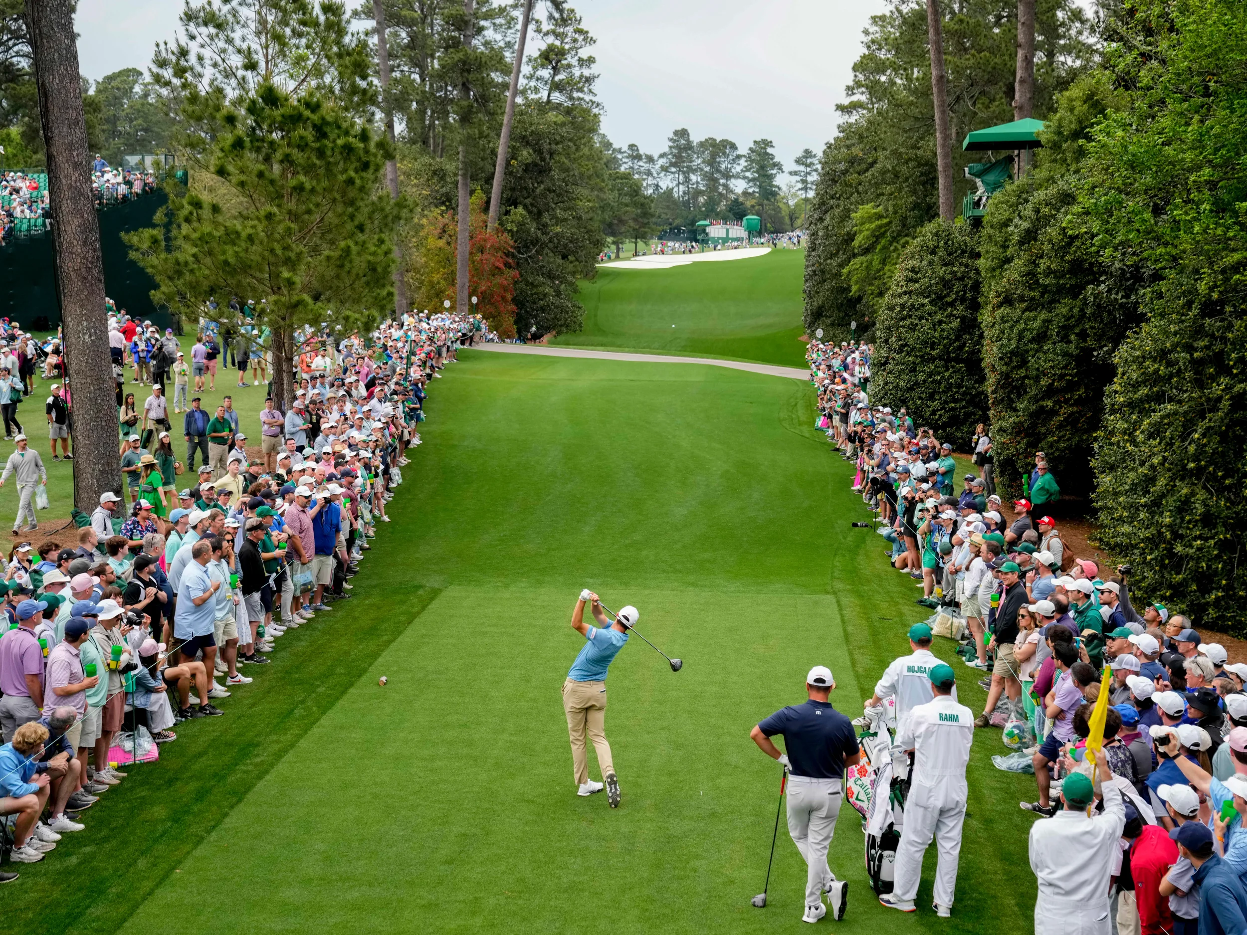 A golfer taking a shot on a lush green golf course lined with spectators on both sides.