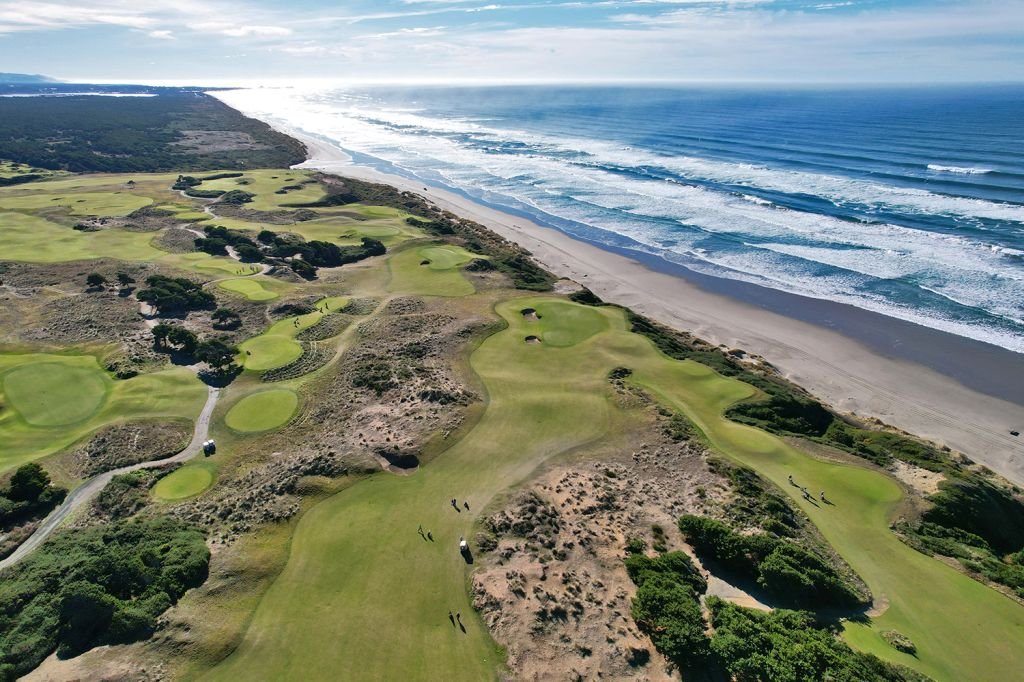 Aerial view of a golf course along a sandy beach with waves crashing onto the shore, under a partly cloudy sky.