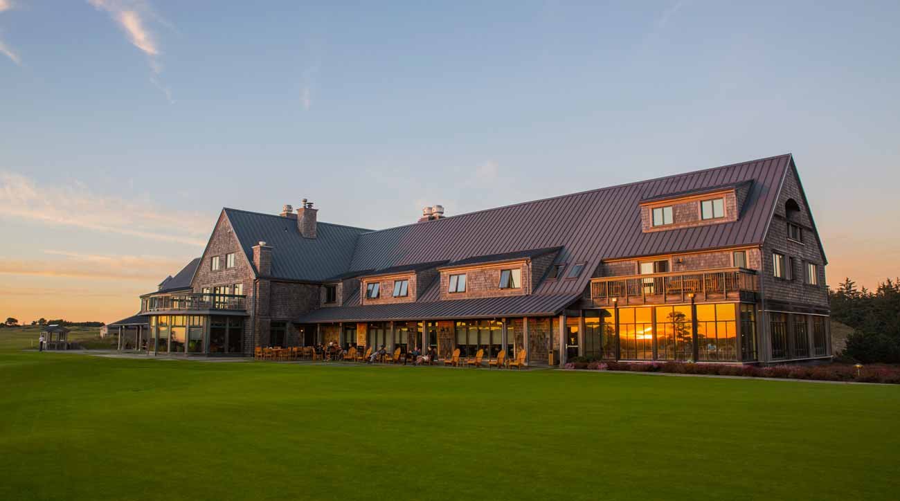 A large house with a dark metal roof and wooden exterior, reflecting the sunset in its glass windows, surrounded by a well-maintained green lawn.