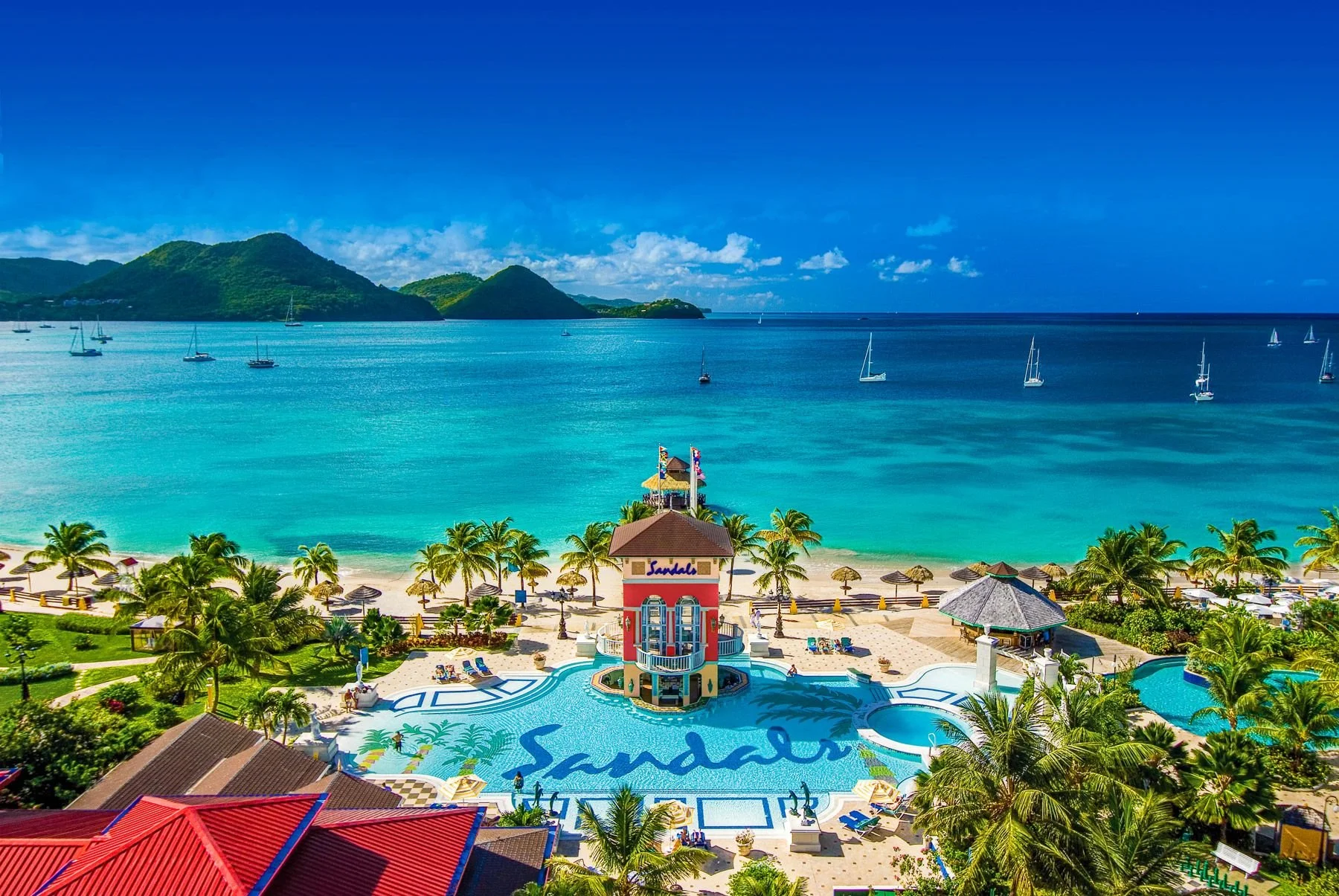 A tropical beach resort with a large pool spelling 'Sandals,' surrounded by palm trees, lounge chairs, and umbrellas, overlooking a clear turquoise ocean with sailboats and green islands in the background on a bright sunny day.