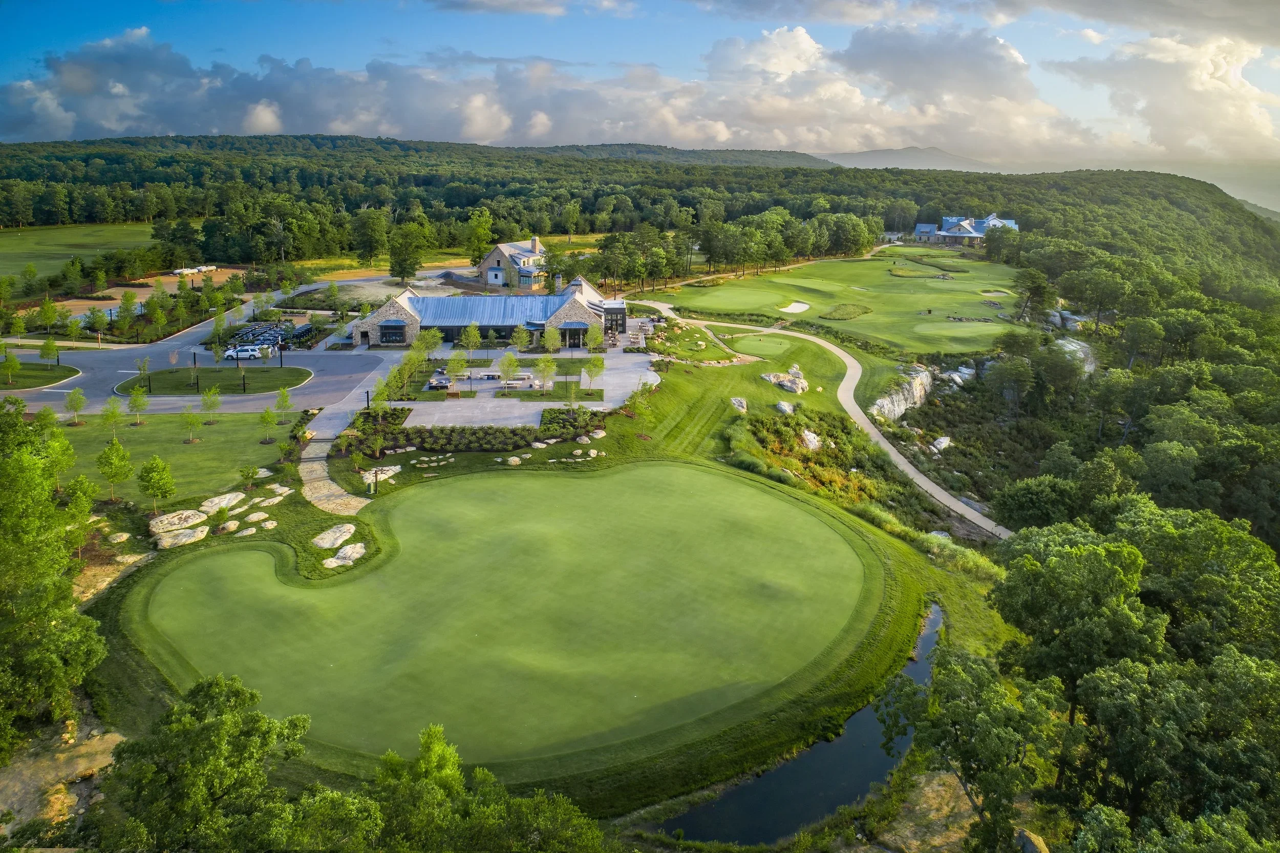 Aerial view of a golf course surrounded by lush green trees, with a clubhouse and parking lot visible in the background, under a partly cloudy sky.