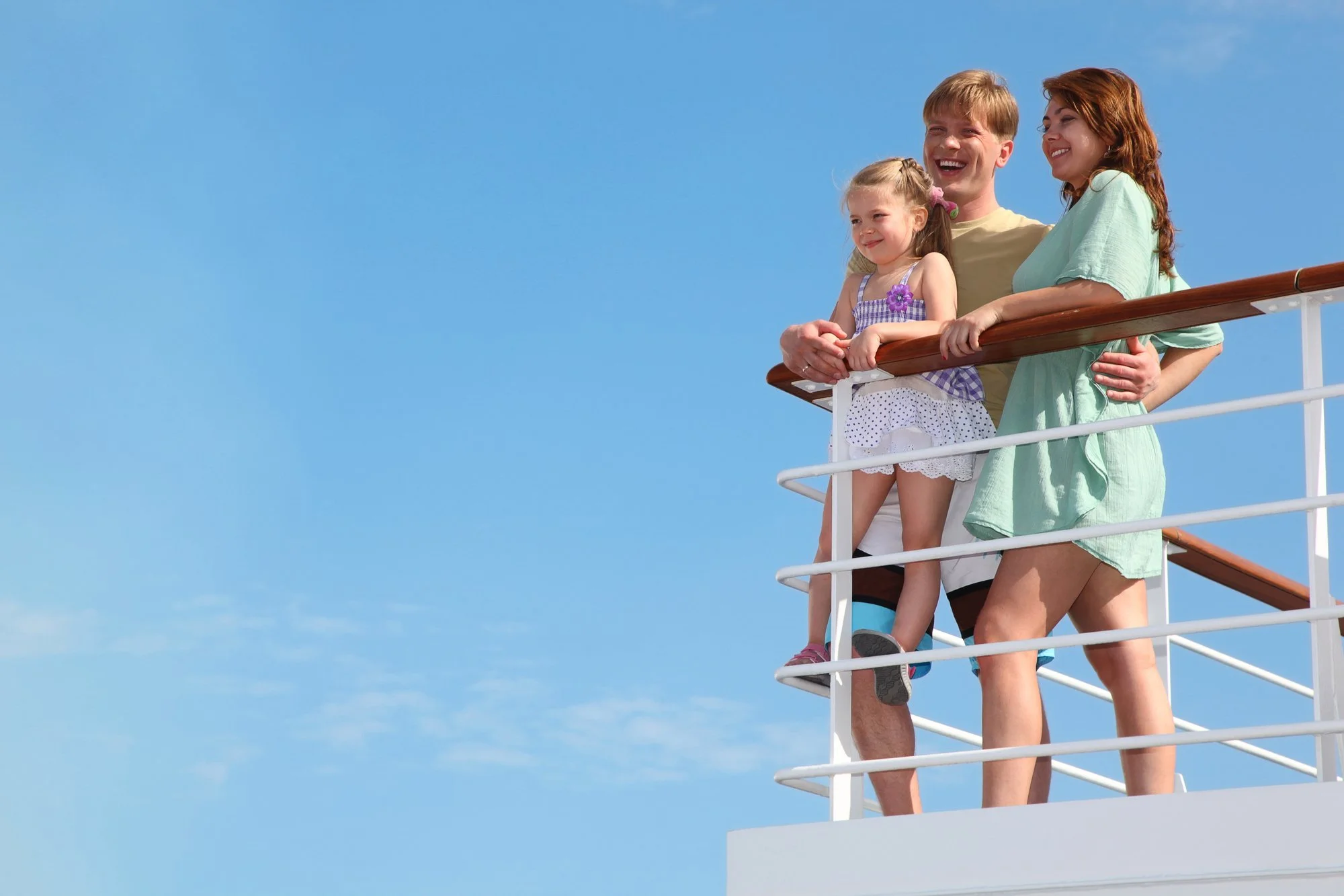 A family of four stands on the deck of a cruise ship, smiling and enjoying a sunny day with a clear blue sky in the background.