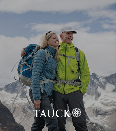 A smiling couple in hiking gear standing on a mountain with snow-covered peaks in the background.
