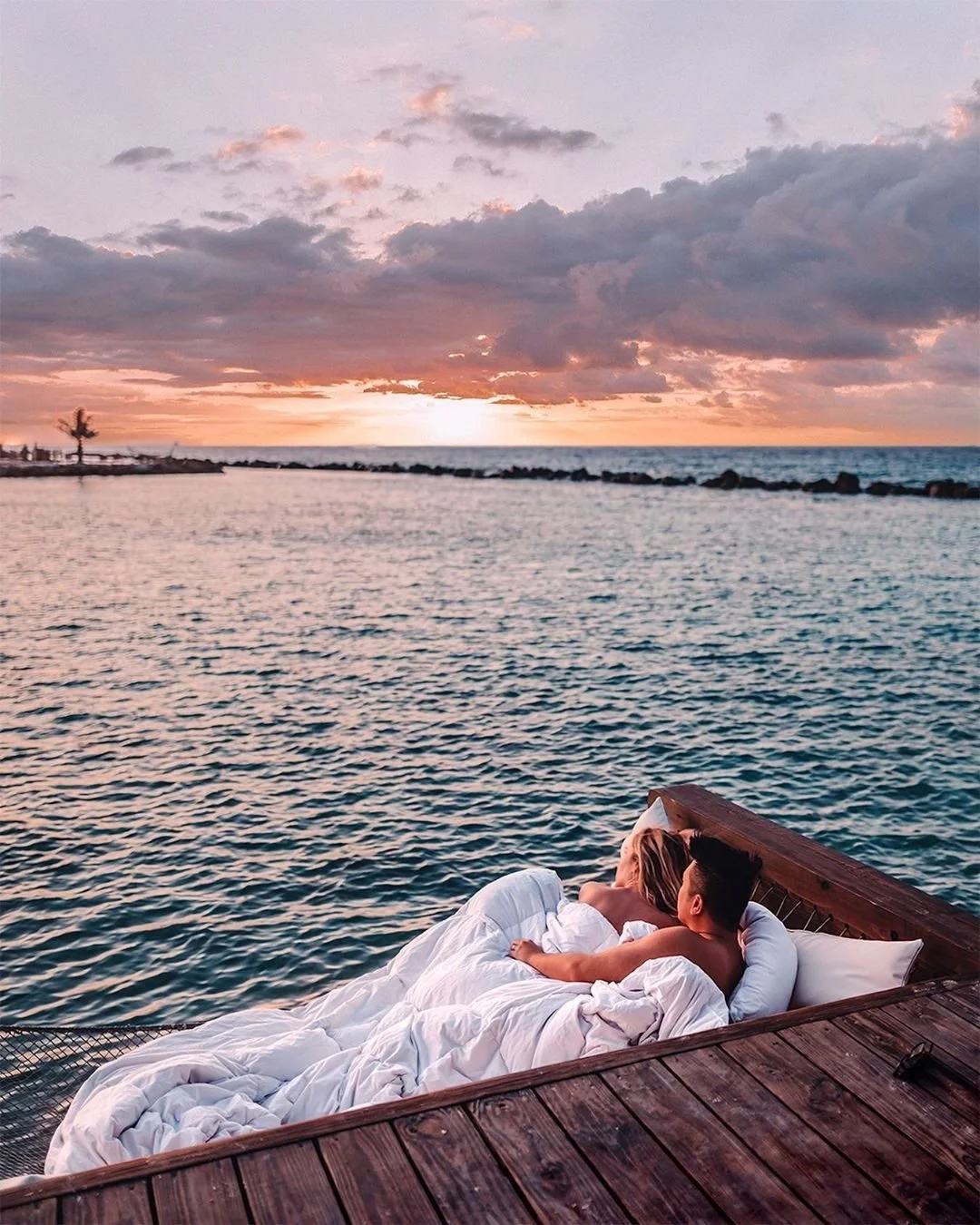 A couple lies on a bed on a wooden dock overlooking the ocean at sunset. The sky is filled with clouds, with the sun setting near the horizon, casting warm light.