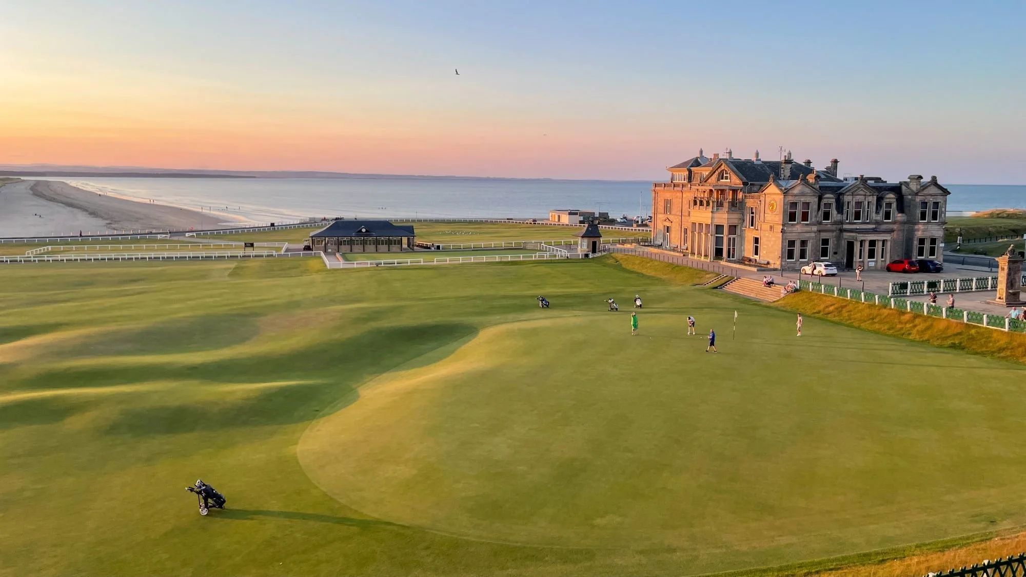 A seaside golf course with a large historic mansion in the background, people playing golf, and a sunset over the ocean.