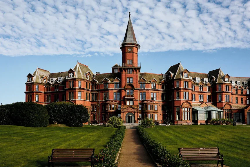 A large historic red brick mansion with a tall turret, surrounded by a well-maintained lawn and a pathway leading to the entrance.