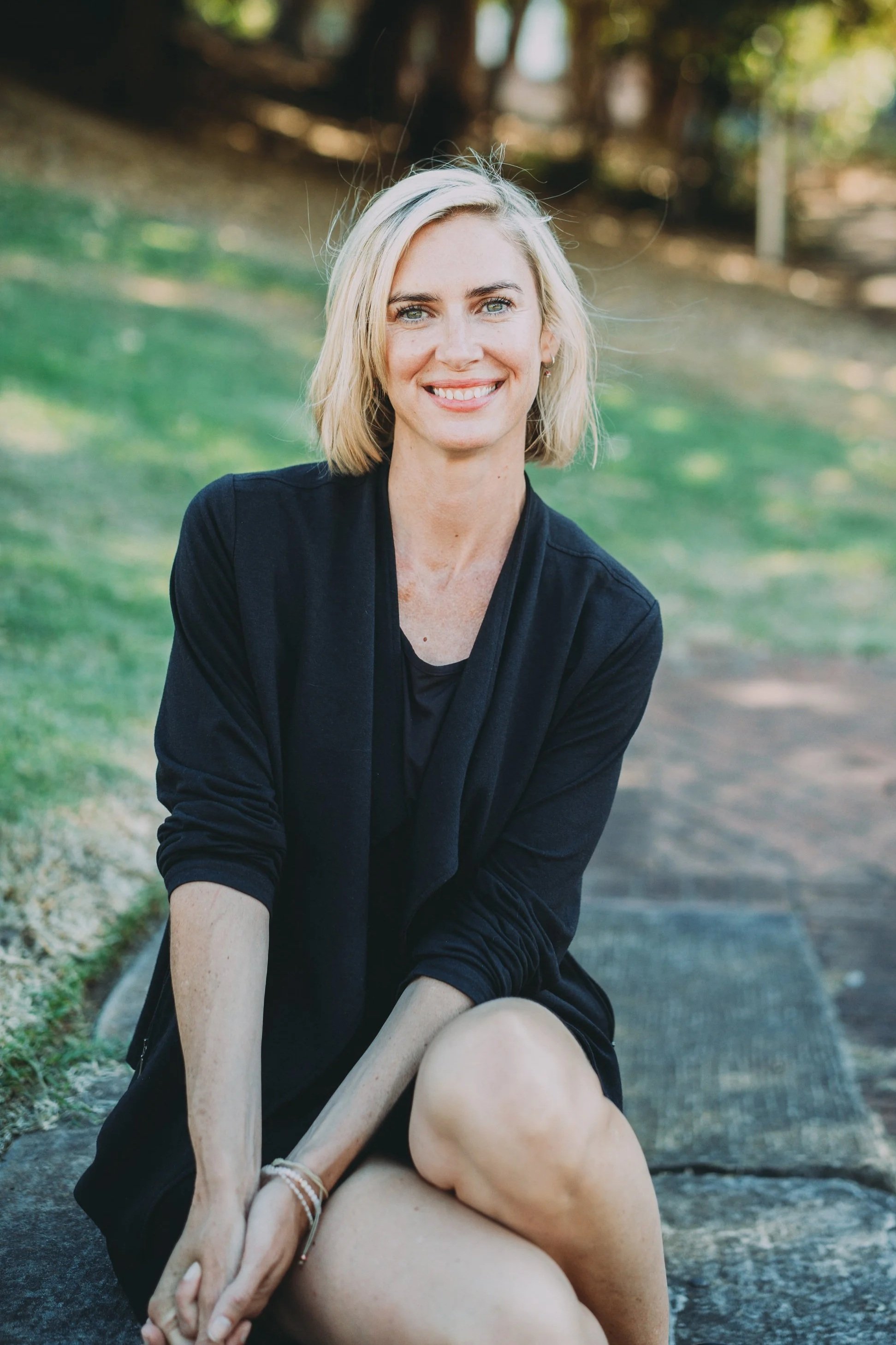 Close-up of a woman with blonde hair, wearing a sleeveless top with red, black, and gold patterns, standing outdoors next to a tree with a textured bark surface.