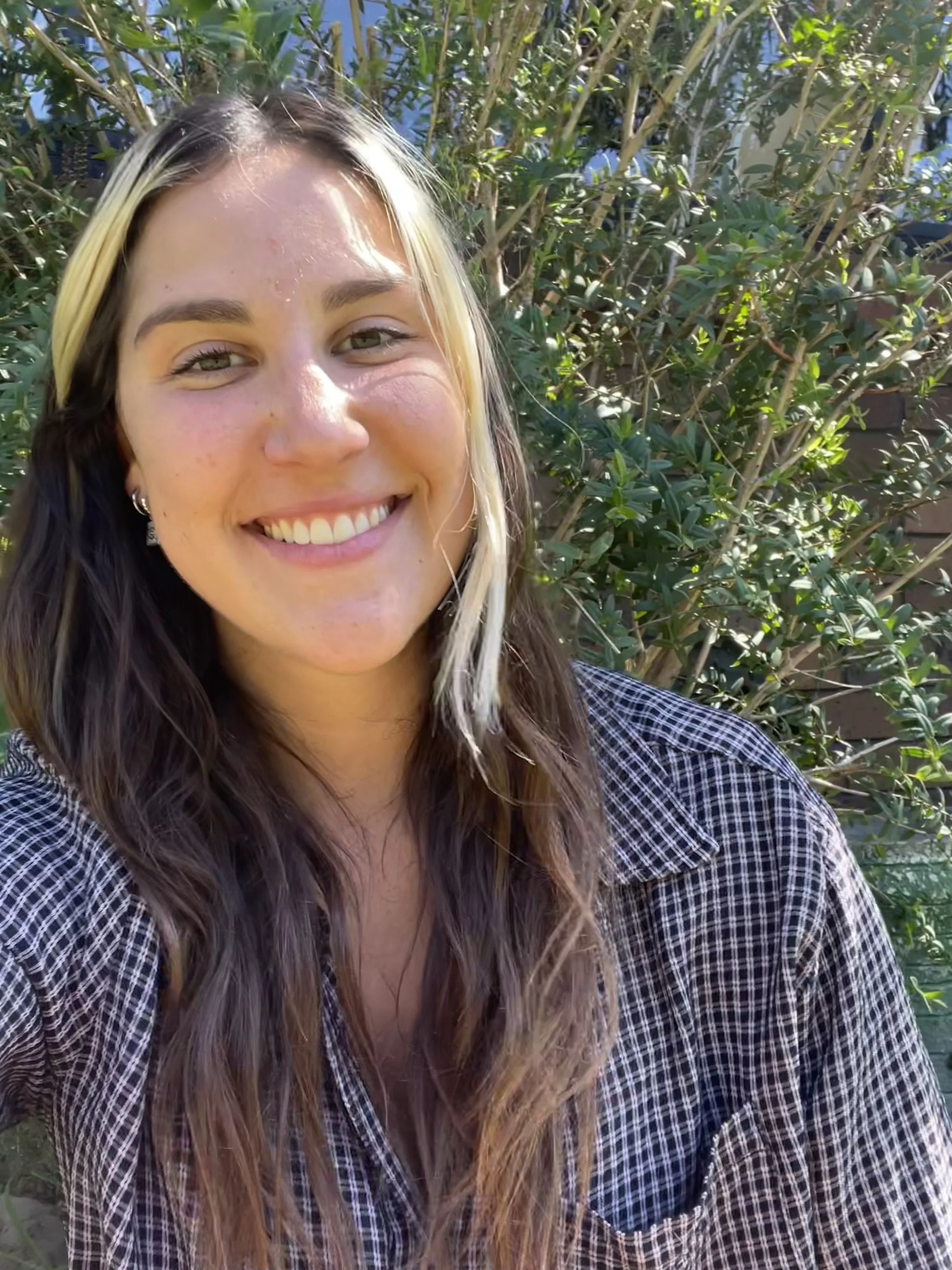Close-up of a young woman smiling outdoors, with greenery in the background, wearing a black and white checkered shirt.