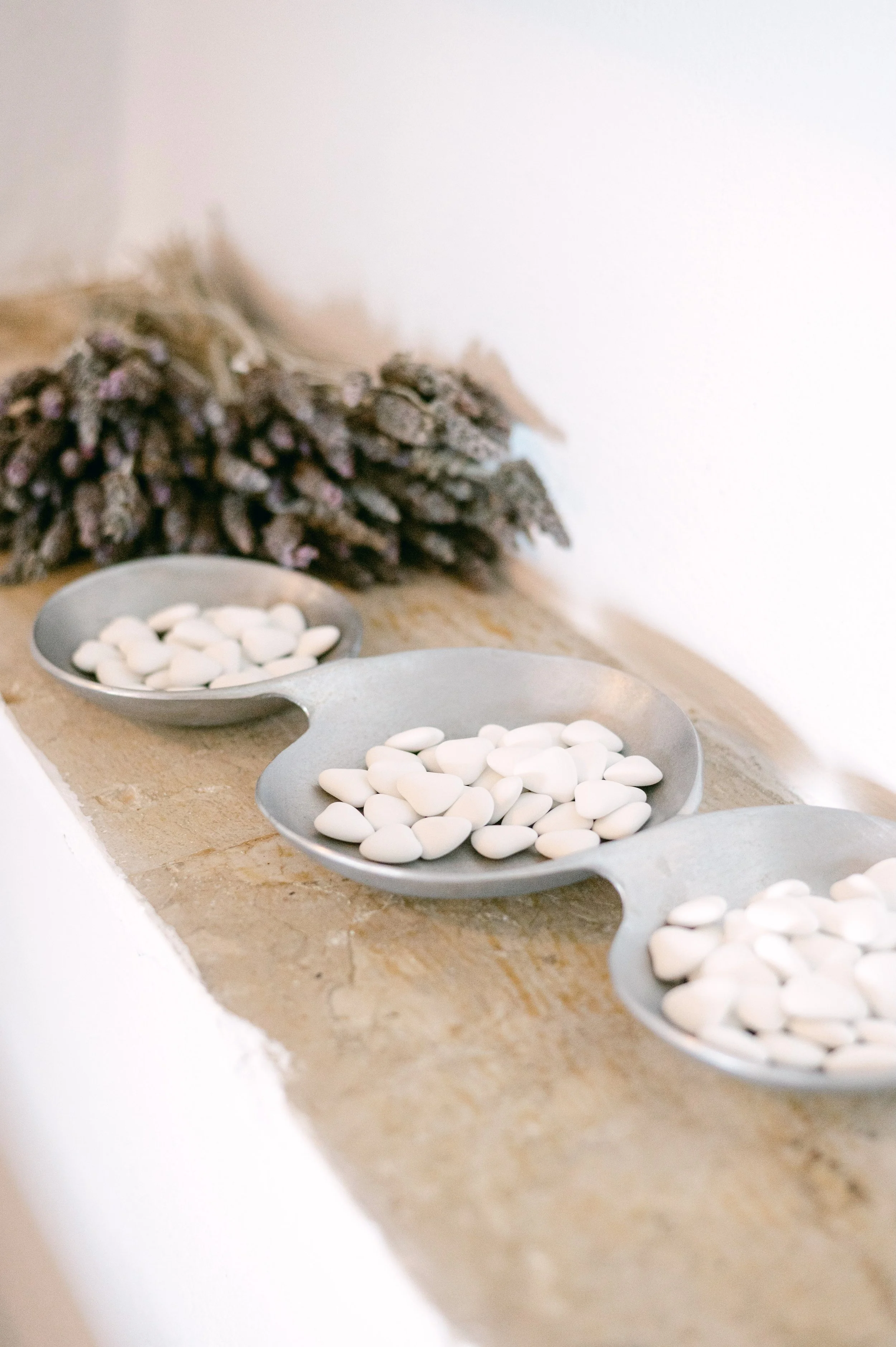 Three metal trays with white almonds placed on a stone surface with a bundle of lavender in the background.