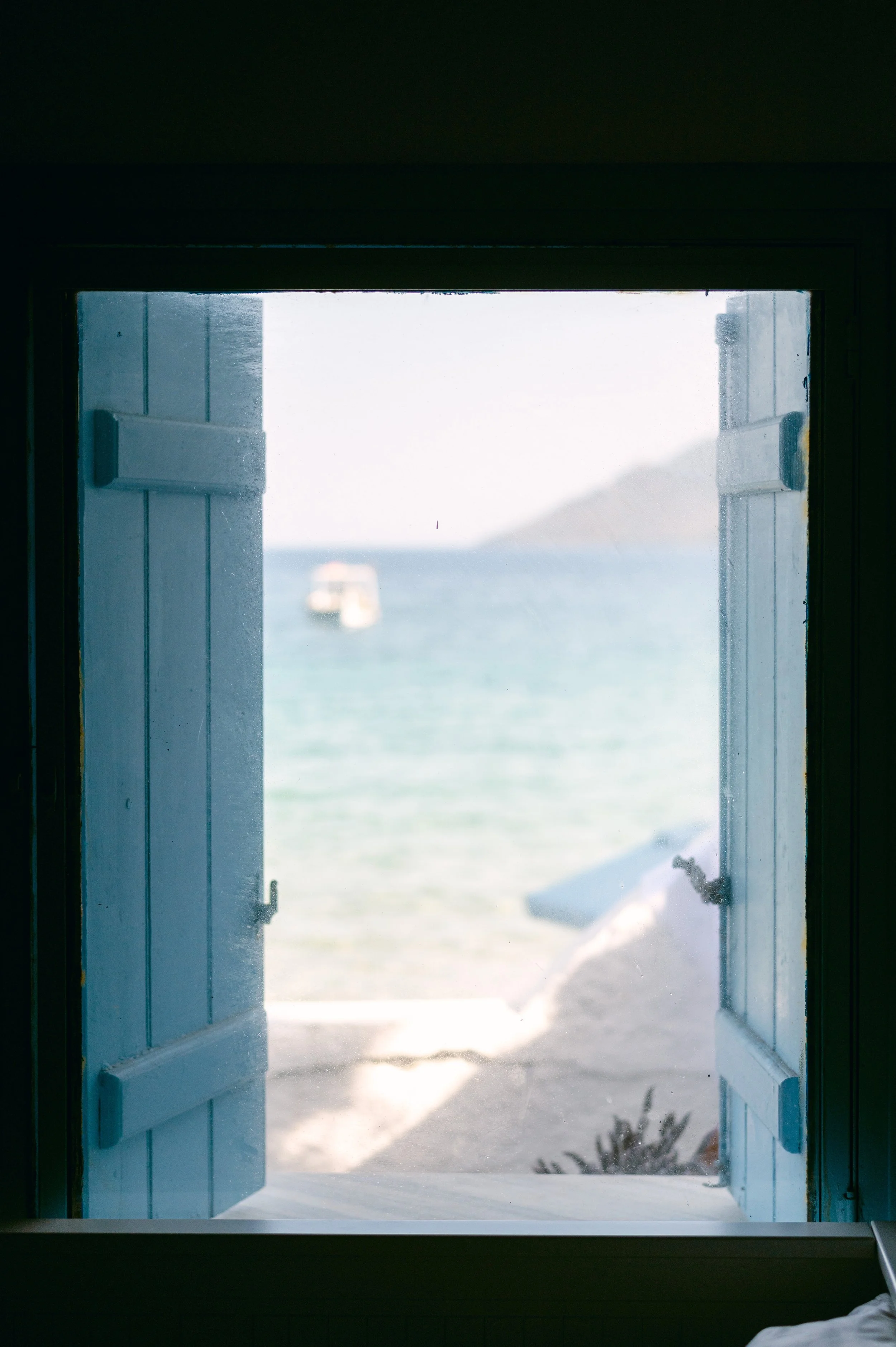 Open window with blue shutters overlooking a beach and ocean, with a boat in the distance and a mountain in the background.