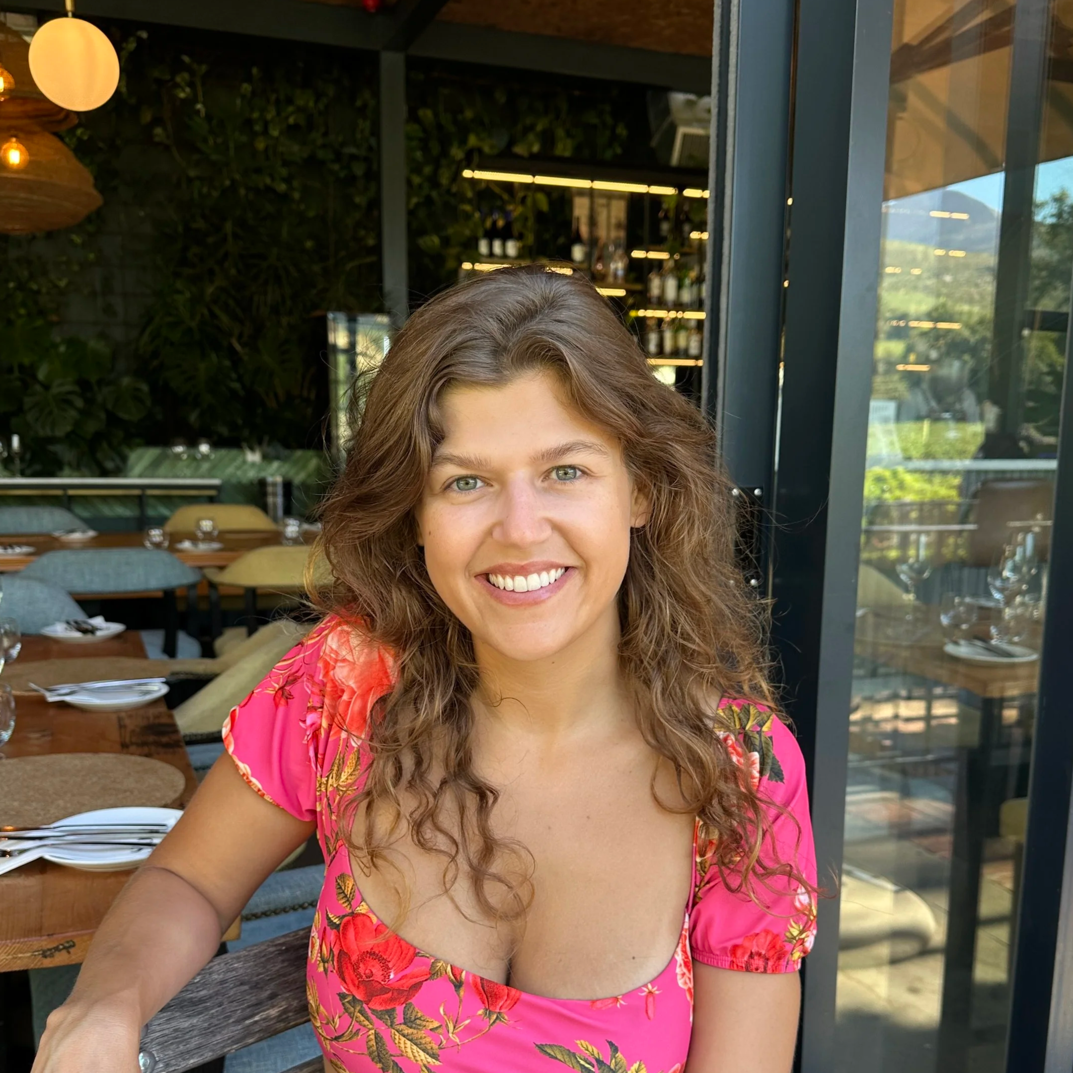 Close-up of a woman smiling with curly brown hair, wearing a pink floral dress, sitting at a dining table in a restaurant with large glass windows, greenery outside, and modern interior decor.