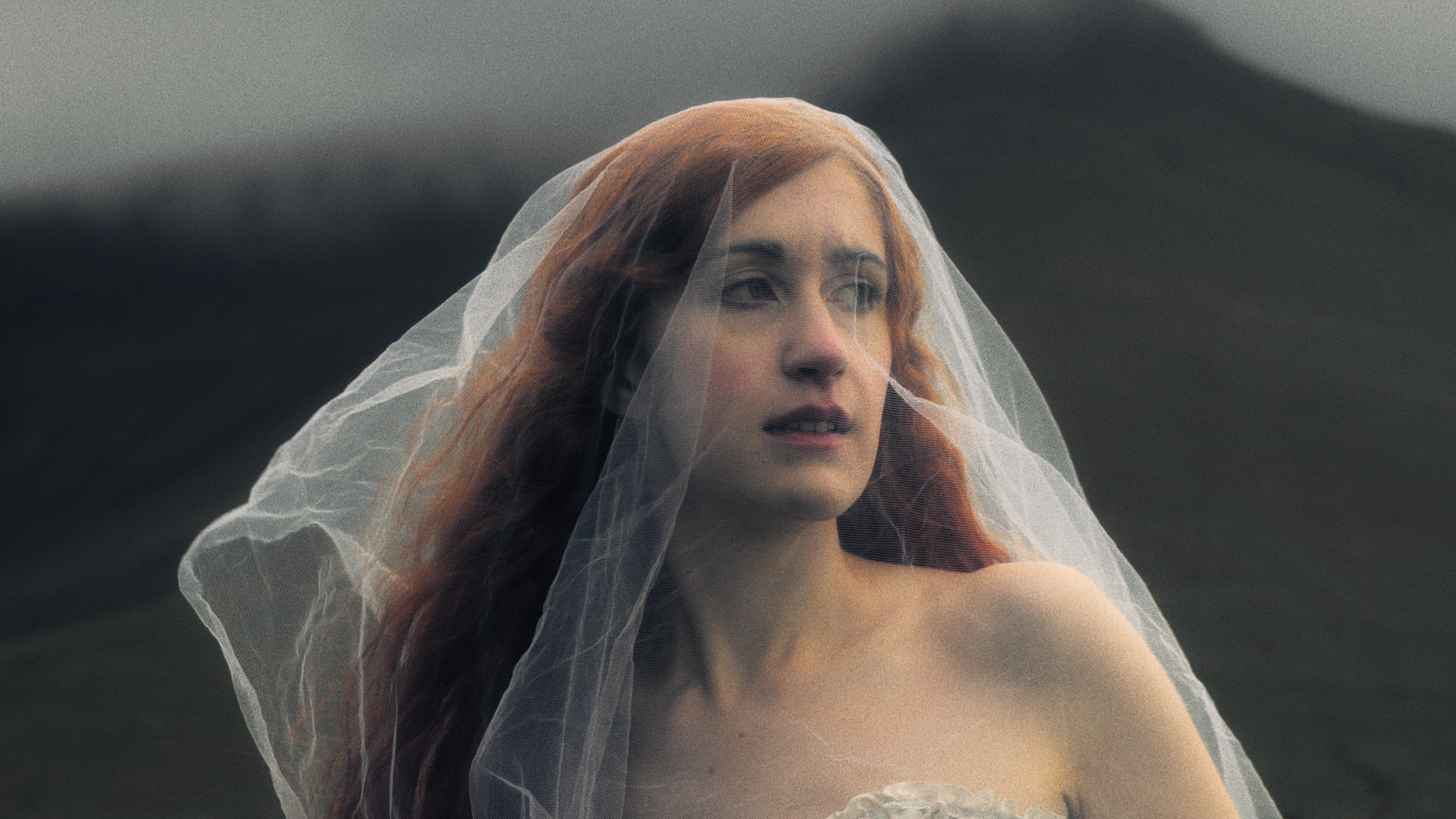 A woman with long red hair, wearing a thin veil, gazing thoughtfully into the distance against a dark, sweeping landscape background.