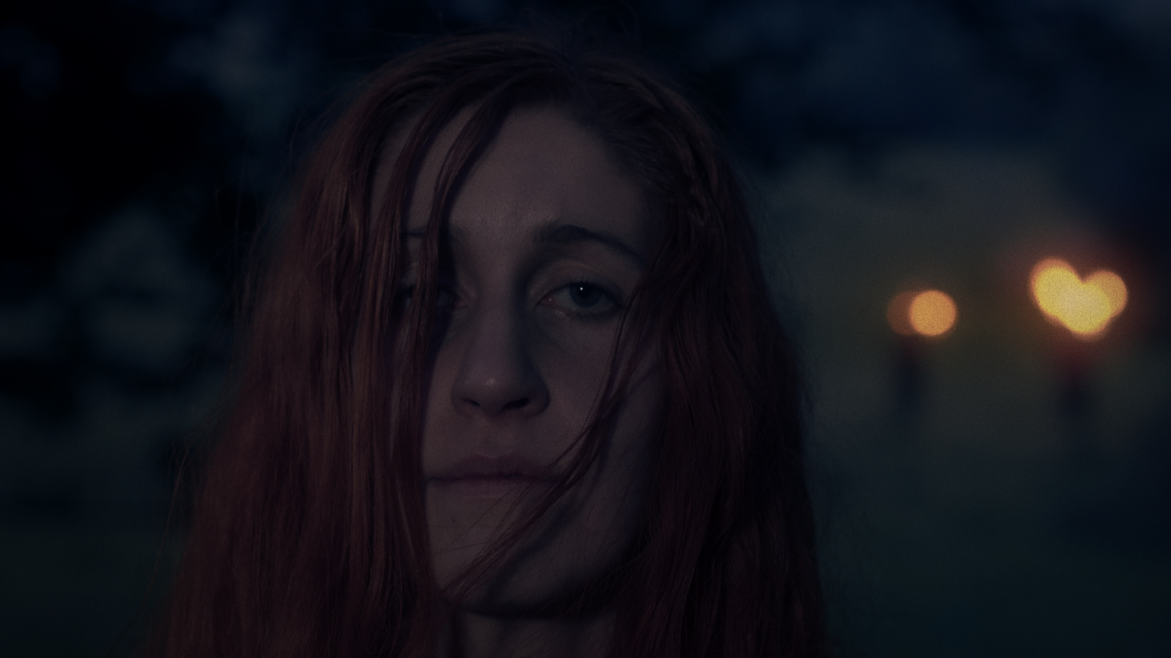 A young woman with red hair standing outdoors during dusk, with blurred lights in the background.