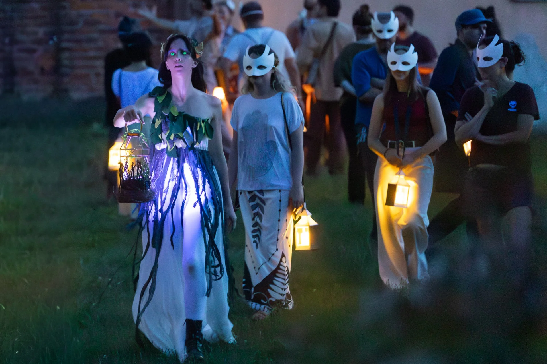 Un'elfa dagli occhi fluorescenti guida con la sua lanterna gli spettatori nel bosco. Gli spettatori indossano una maschera bianca che gli copre parzialmente il volto. Ogni spettatore ha una lanterna.

Foto di Davide Aiello.