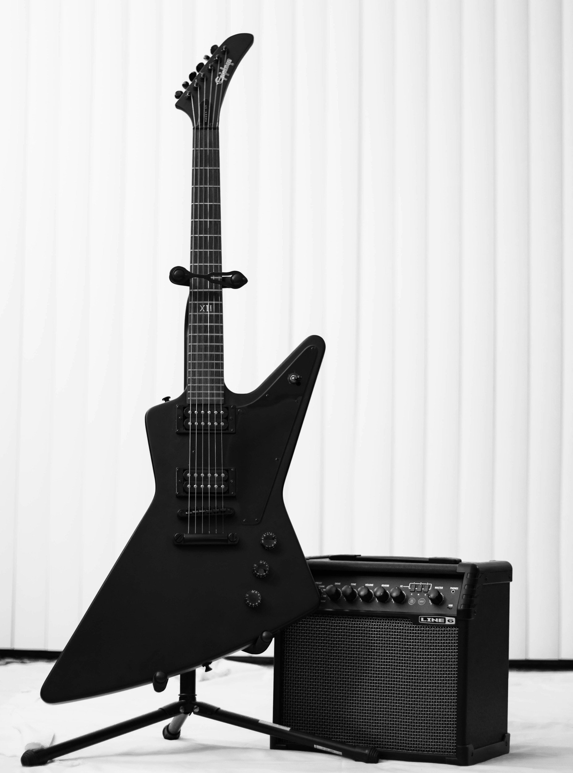 A black electric guitar with a unique, angular shape, placed on a stand next to a small guitar amplifier, with a white vertical panel background.