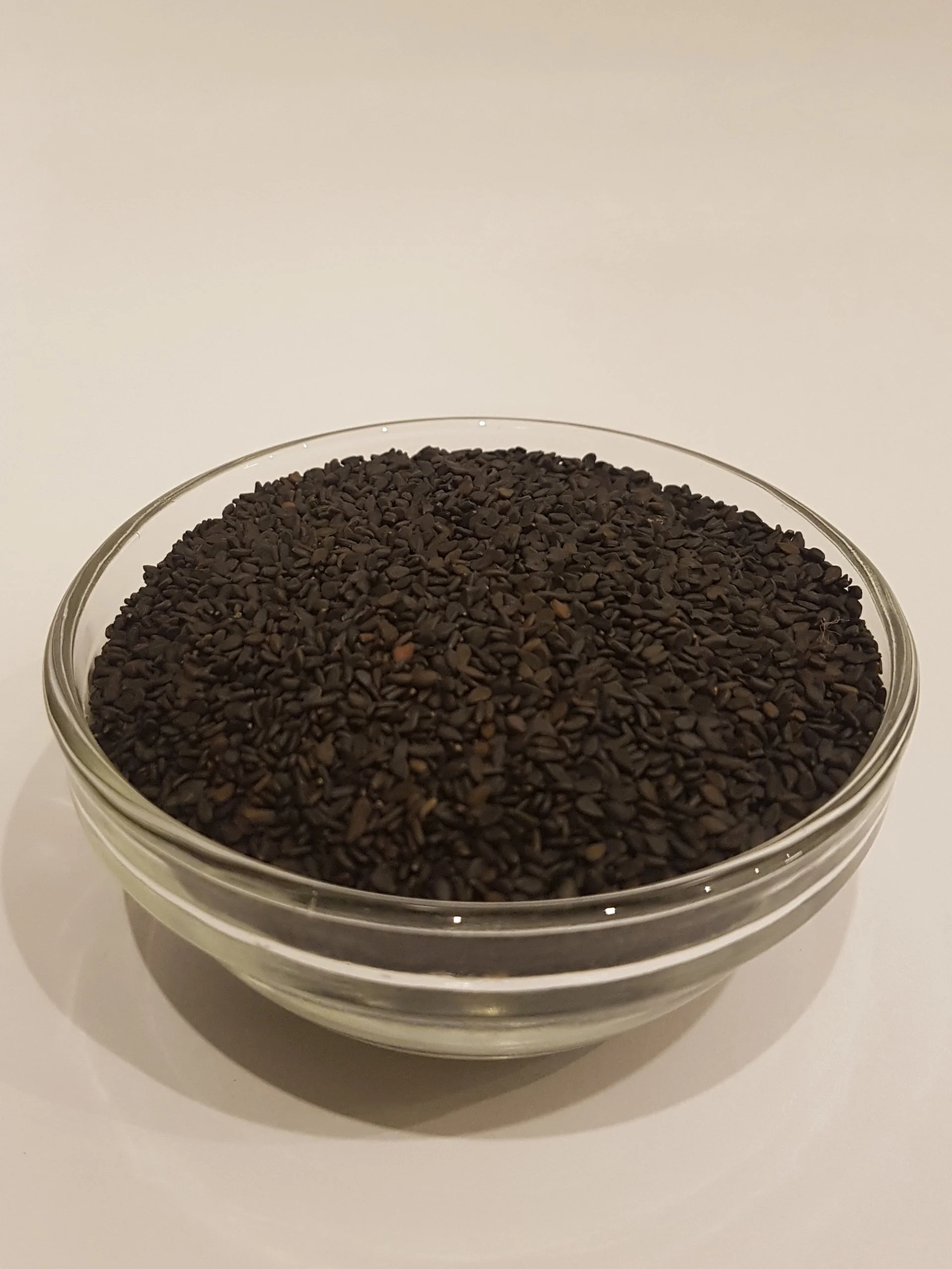 A glass bowl filled with small black sesame seeds on a white surface.