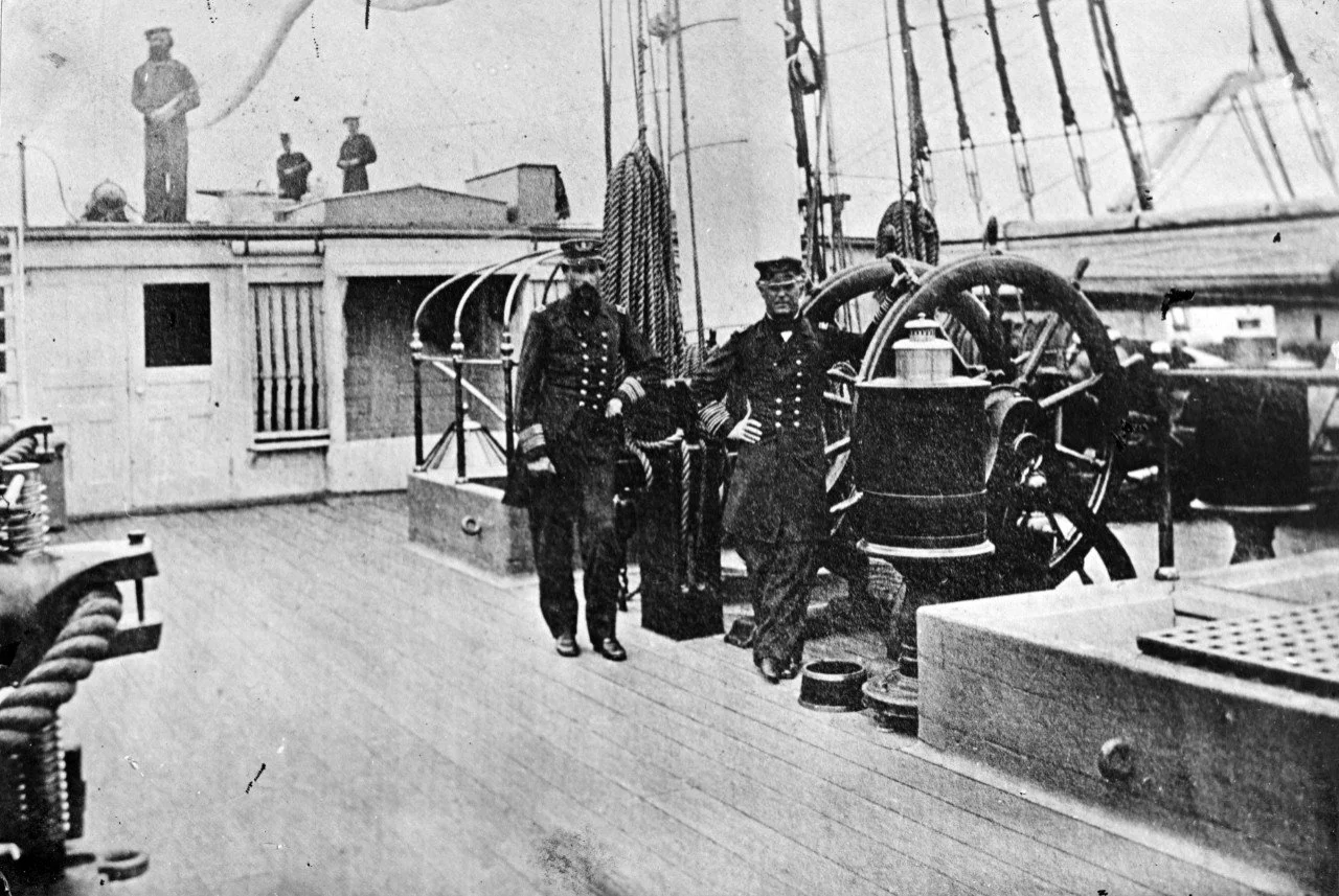 Photo taken aboard USS Hartford in 1864.  Admiral David Farragut and Captain Percival Drayton stand beside the ship’s wheel.  The breech of 30-Pounder Parrott may be see on the poop deck at the upper left of the photo.