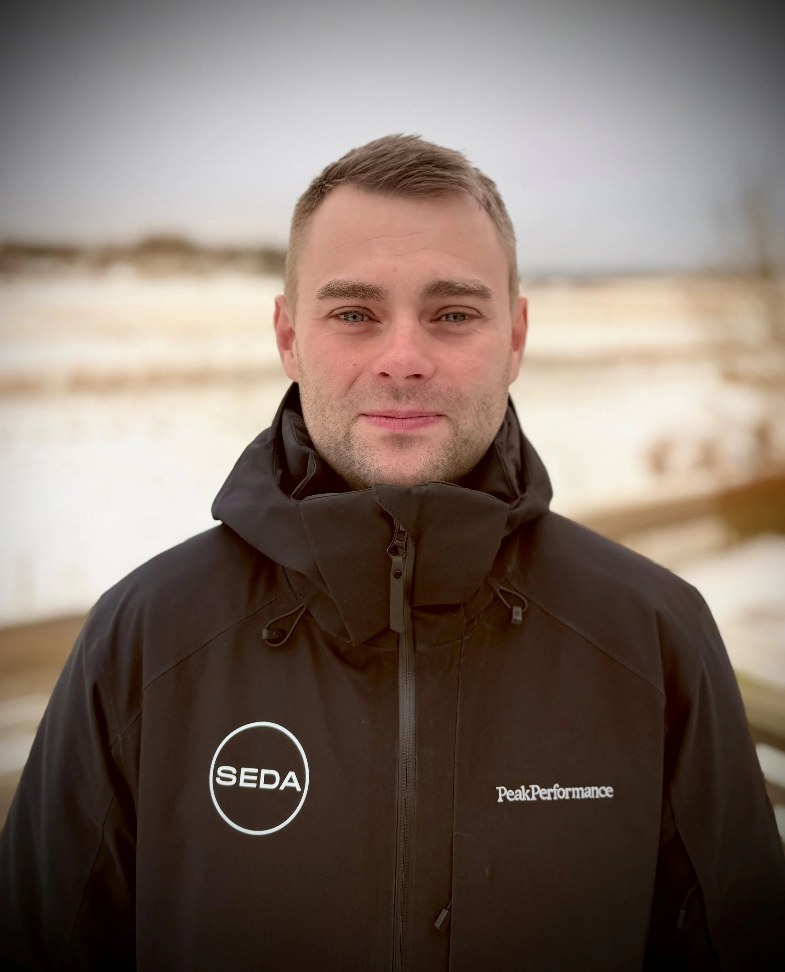 A man in a black hooded jacket outdoors with a blurred snowy landscape in the background.