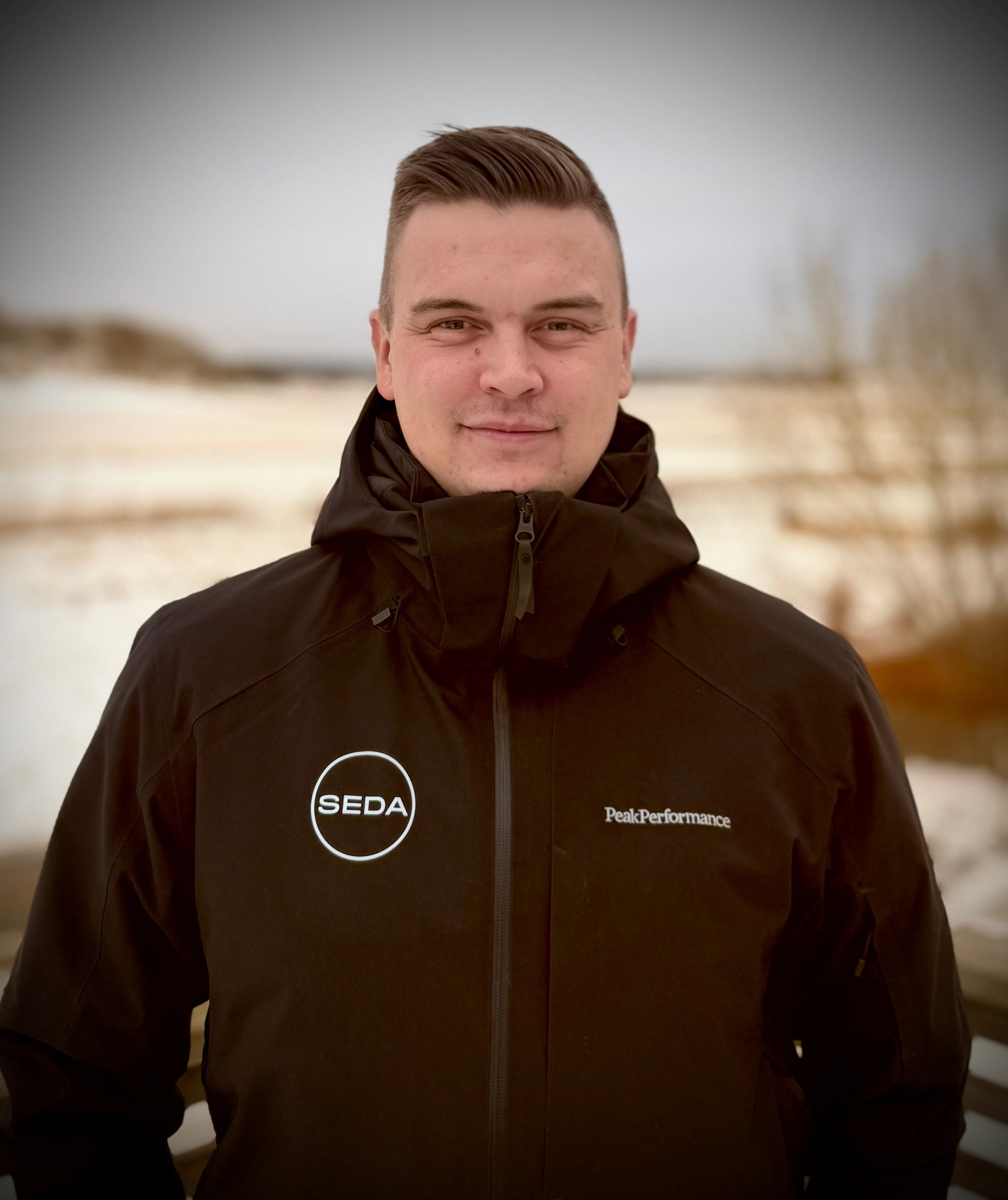 A young man with short brown hair wearing a black jacket outdoors in a snowy landscape.