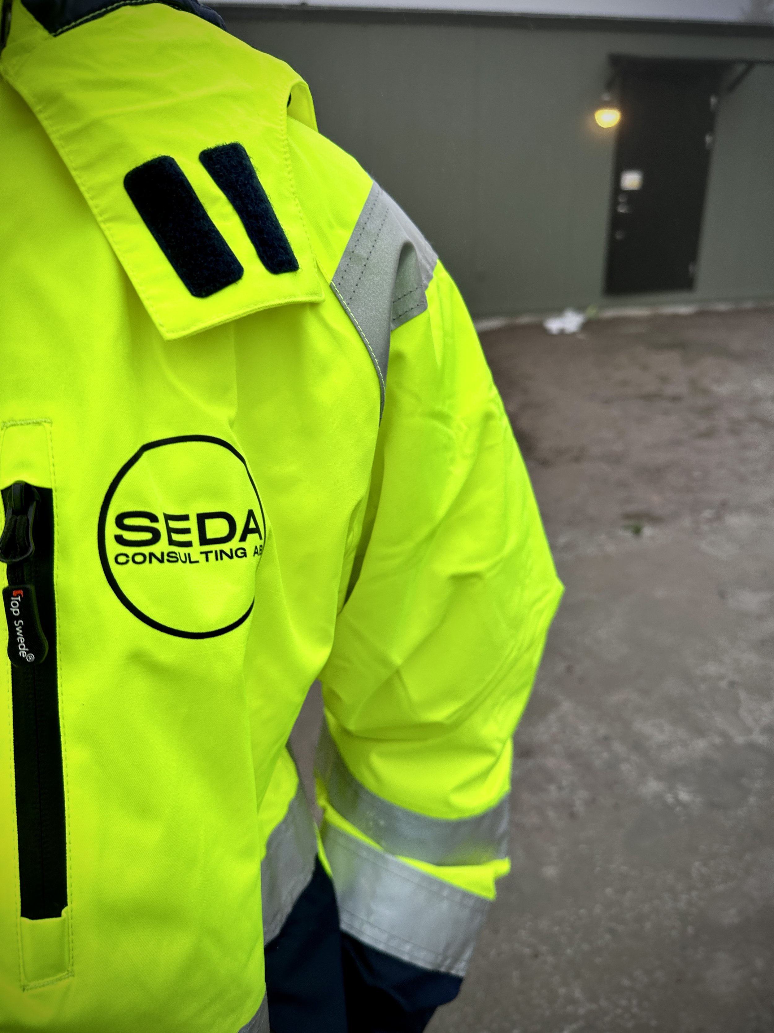 Close-up of a person wearing a bright yellow safety jacket with the logo 'SEDA Consulting' on the chest, standing on a concrete ground near a dark gray wall with a small door and some scattered debris.