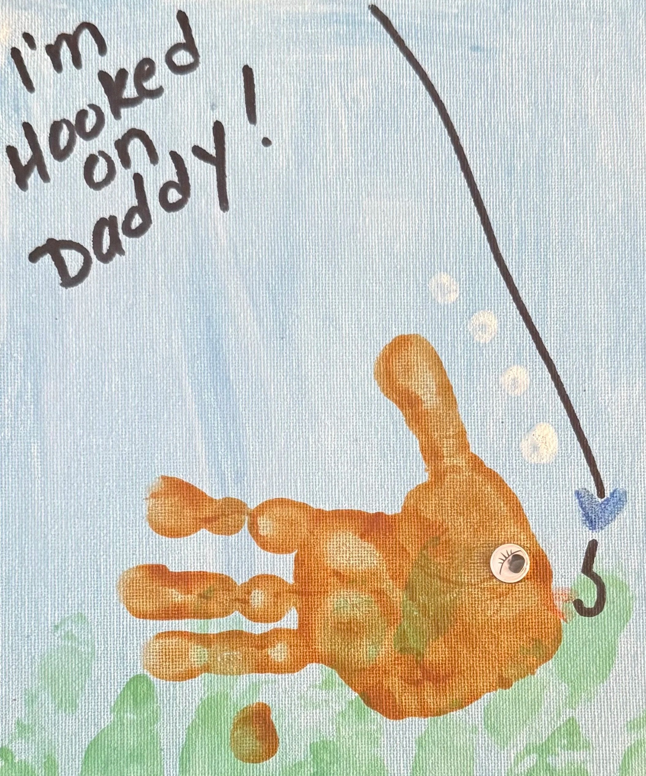 Child's drawing of an orange fish with a googly eye, green fins, and a black fishing hook with a blue and purple line, surrounded by green grass and a light blue sky, with handwritten note saying 'I'm hooked on Daddy!'.
