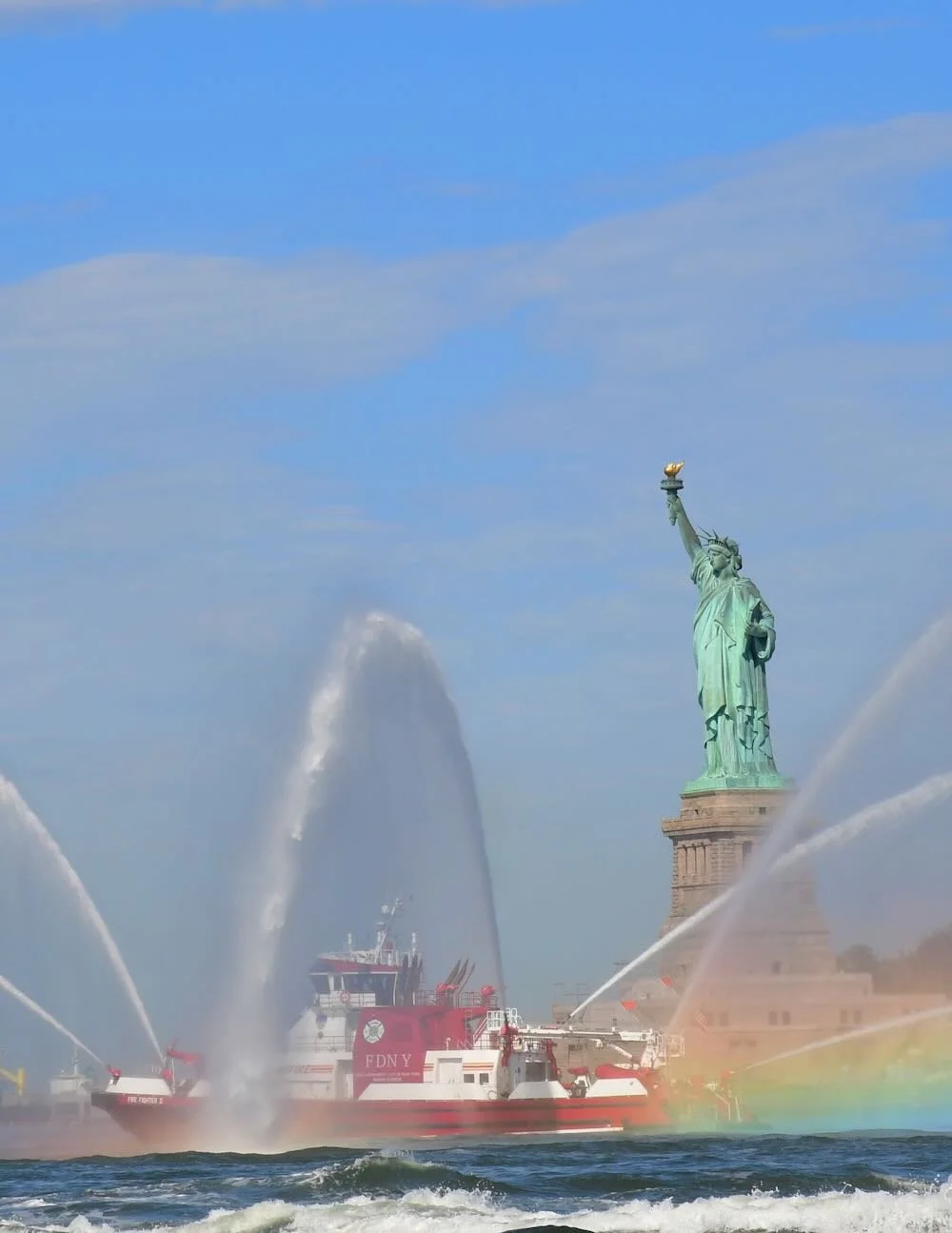 statue of liberty with fireboat shooting water out with blue skies and blue waters