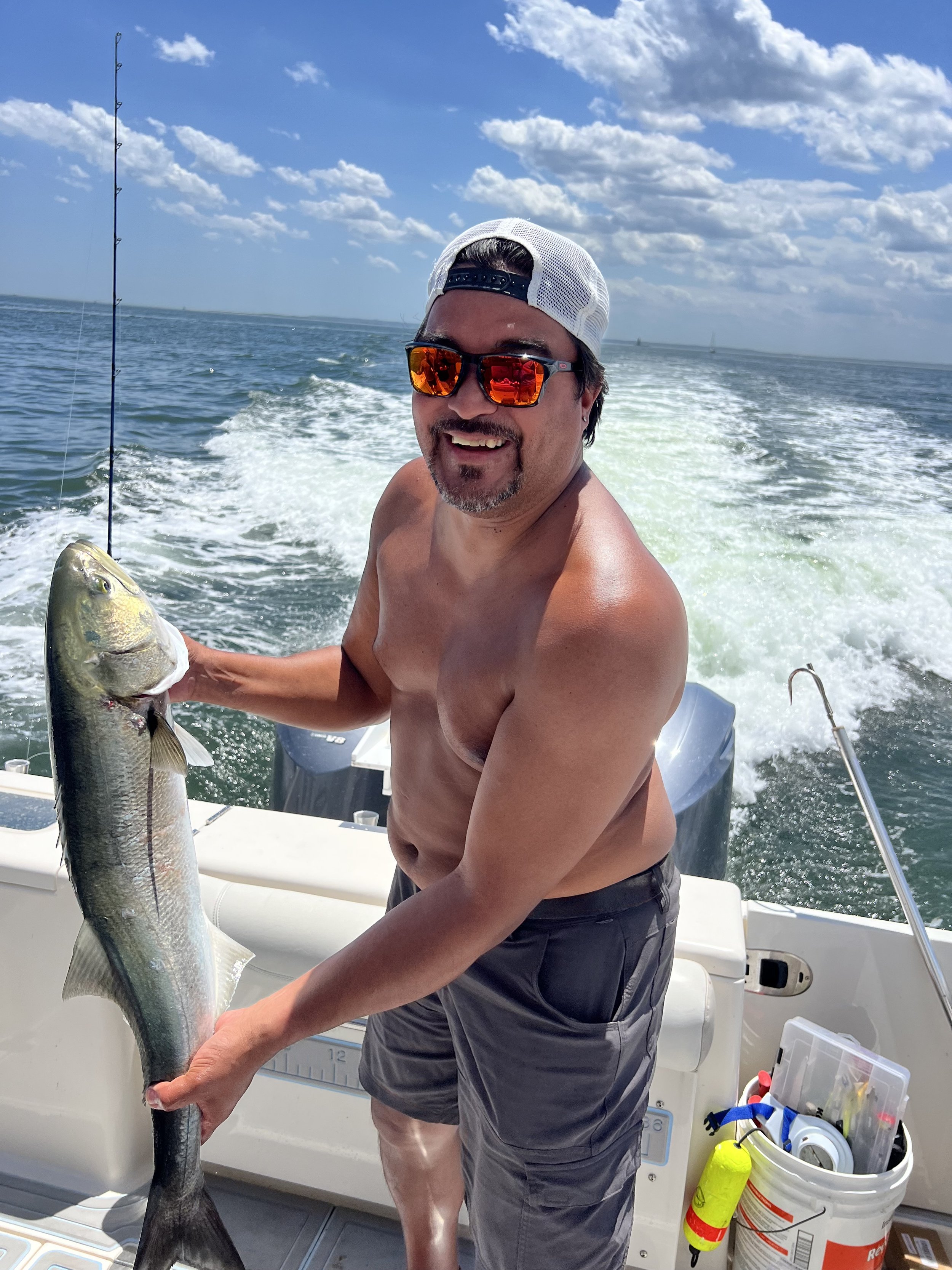 man with sunglasses and backwards hat holding a blue fish on a boat with blue skies and puffy clouds and ocean behin