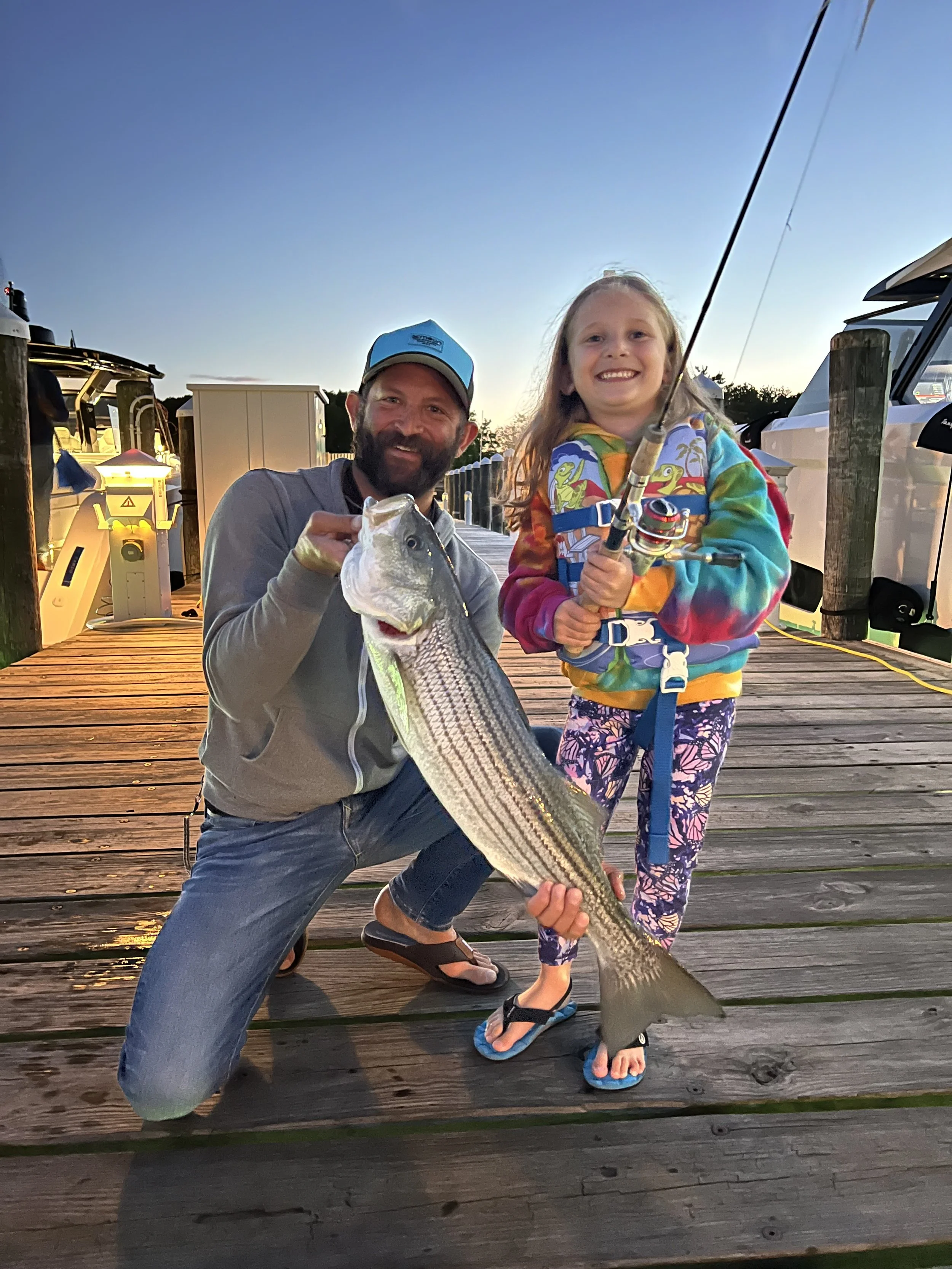 A man and a young girl on a wooden dock holding a large striped bass fish, smiling, with boats and sky in the background during sunset.