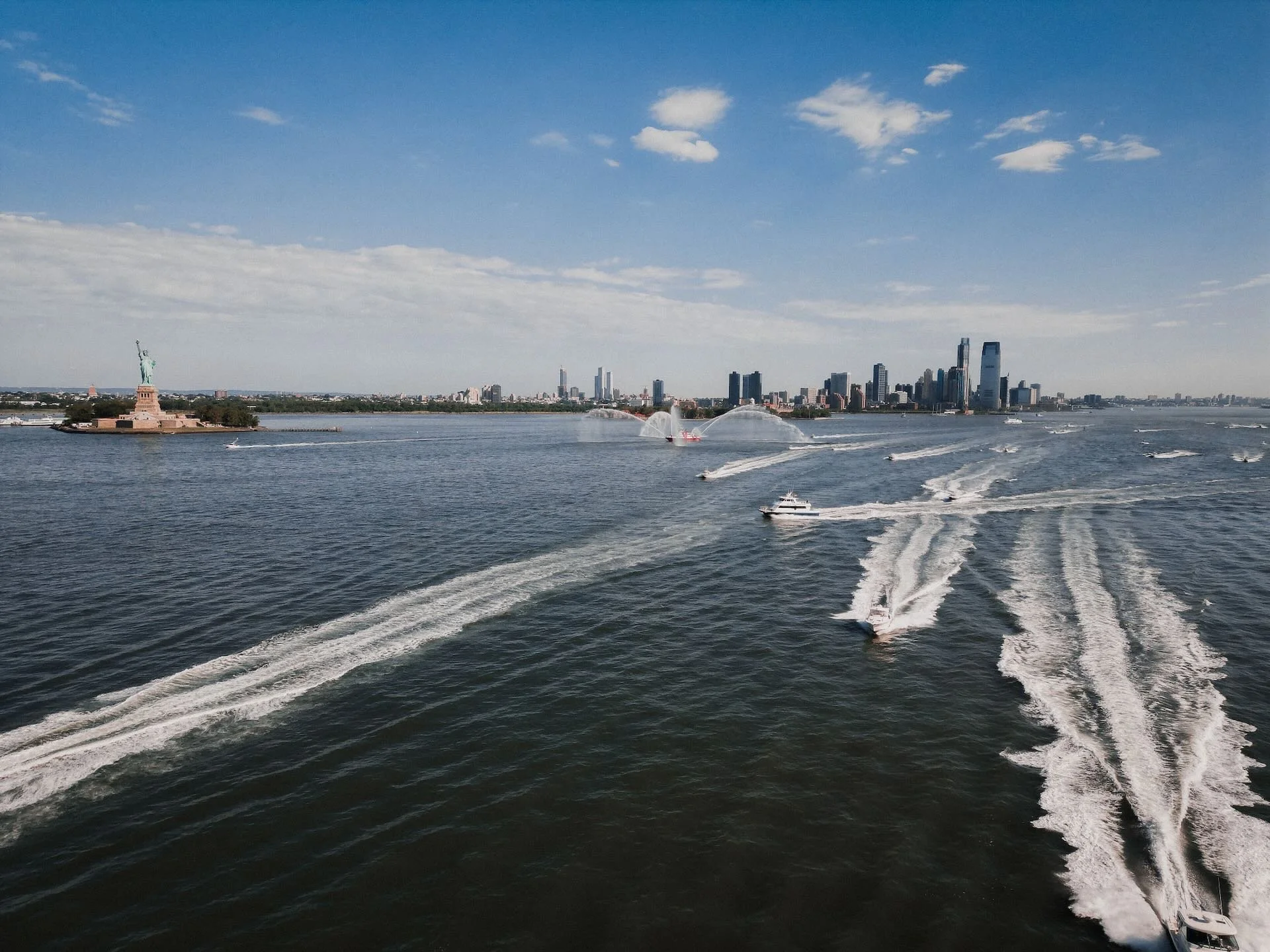 NY harbor with view of NYC and statue of liberty with many boats speeding through the water on a sunny day with some clouds