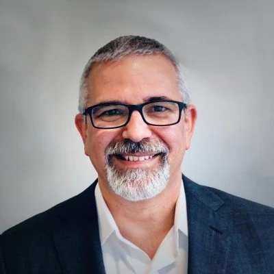 Portrait of a middle-aged man with glasses, short gray hair, and a beard, wearing a dark suit and white shirt, smiling against a plain background.