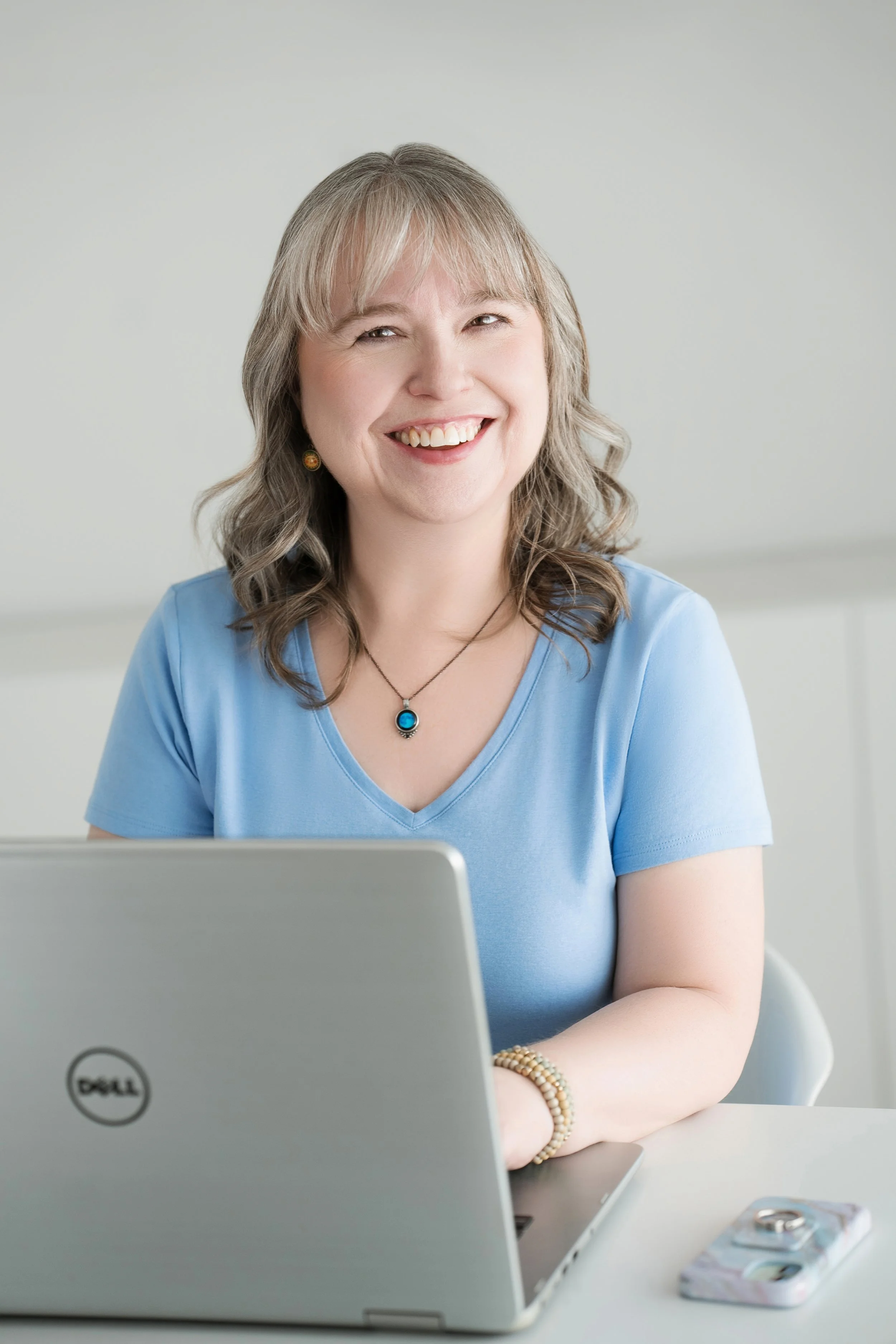 A smiling woman, Dawn Josephson, with shoulder-length wavy hair wearing a light blue t-shirt, sitting at a desk with a Dell laptop and a smartphone with a marble case.